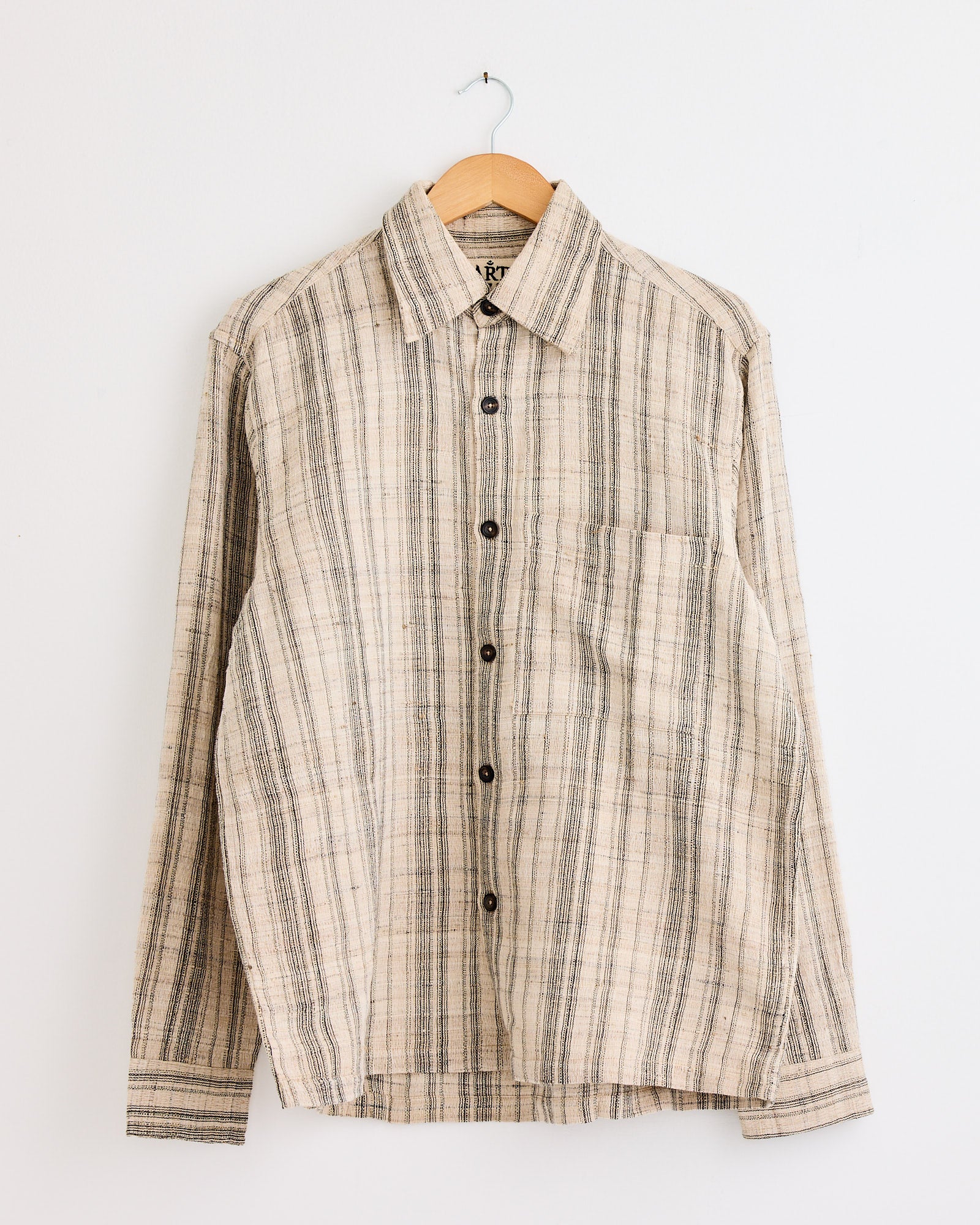 The Box Striped Shirt in Black/Ecru by Kartik Research, featuring black and ecru vertical stripes and long sleeves, hangs on a wooden hanger against a white wall.