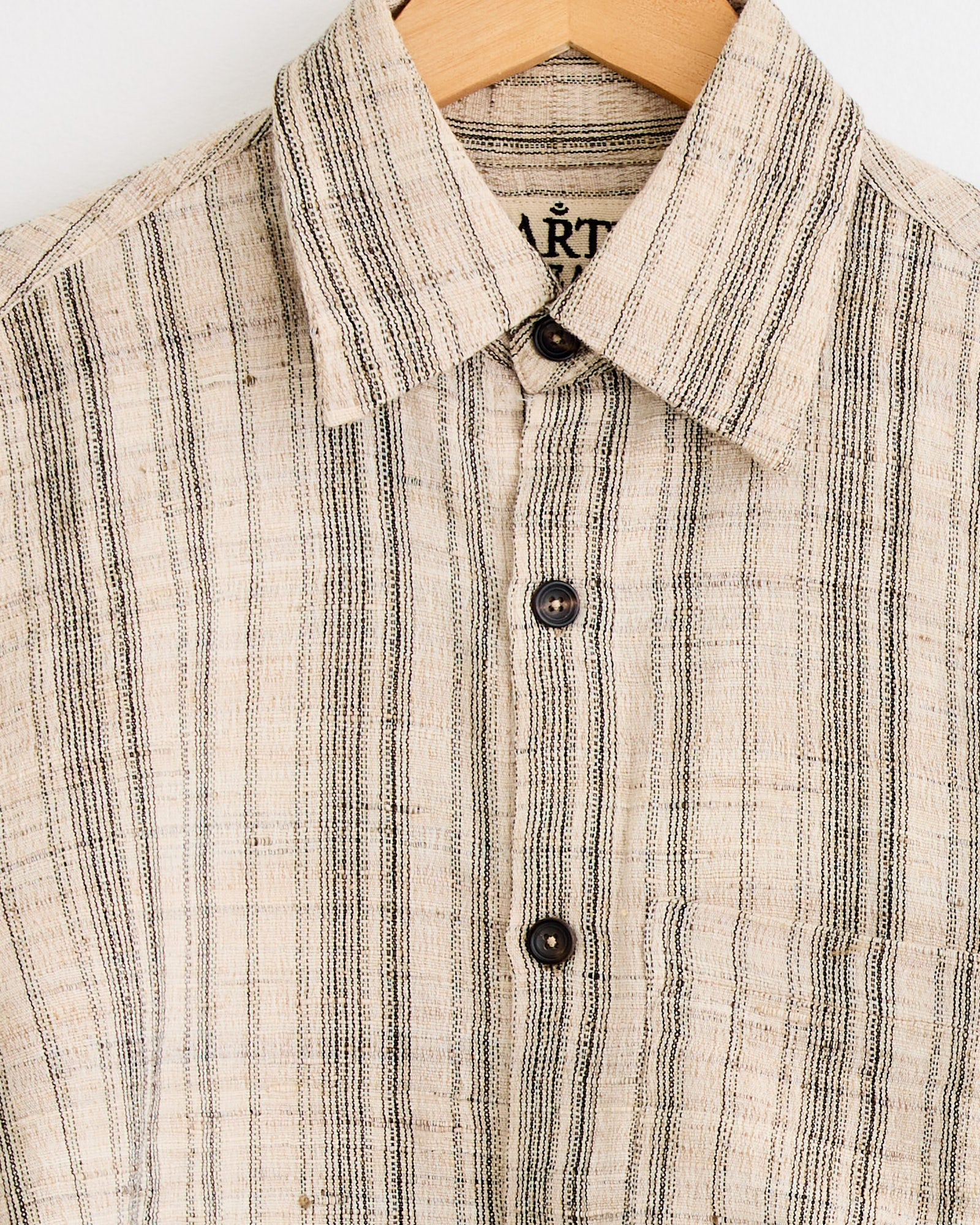 Close-up of the Kartik Research Box Striped Shirt in Black/Ecru, featuring vertical stripes, a collar, and three dark buttons. This hand-embroidered cotton shirt hangs on a wooden hanger against a plain background.