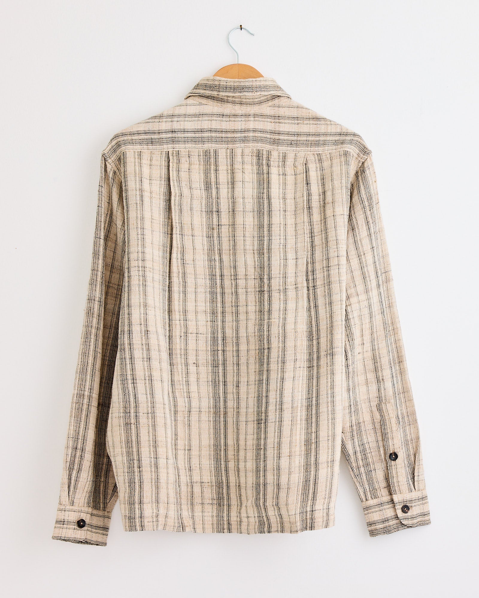 A Kartik Research Box Striped Shirt in Black/Ecru hangs on a wooden hanger against a white wall, shown from the back. This hand-embroidered cotton shirt features bold vertical stripes for a modern look.