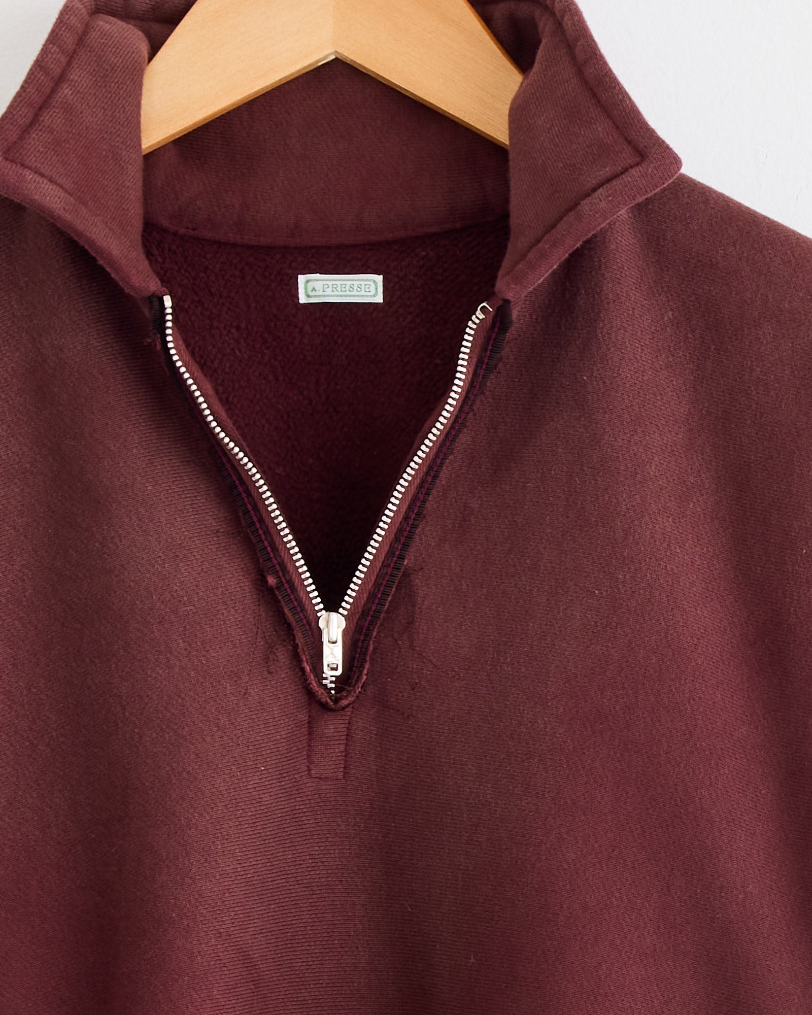 A close-up of the A.PRESSE Vintage Half Zip Sweatshirt in Burgundy hangs on a wooden hanger with its zipper slightly open, revealing a soft lining. For sizing or fit questions, contact us via online chat.