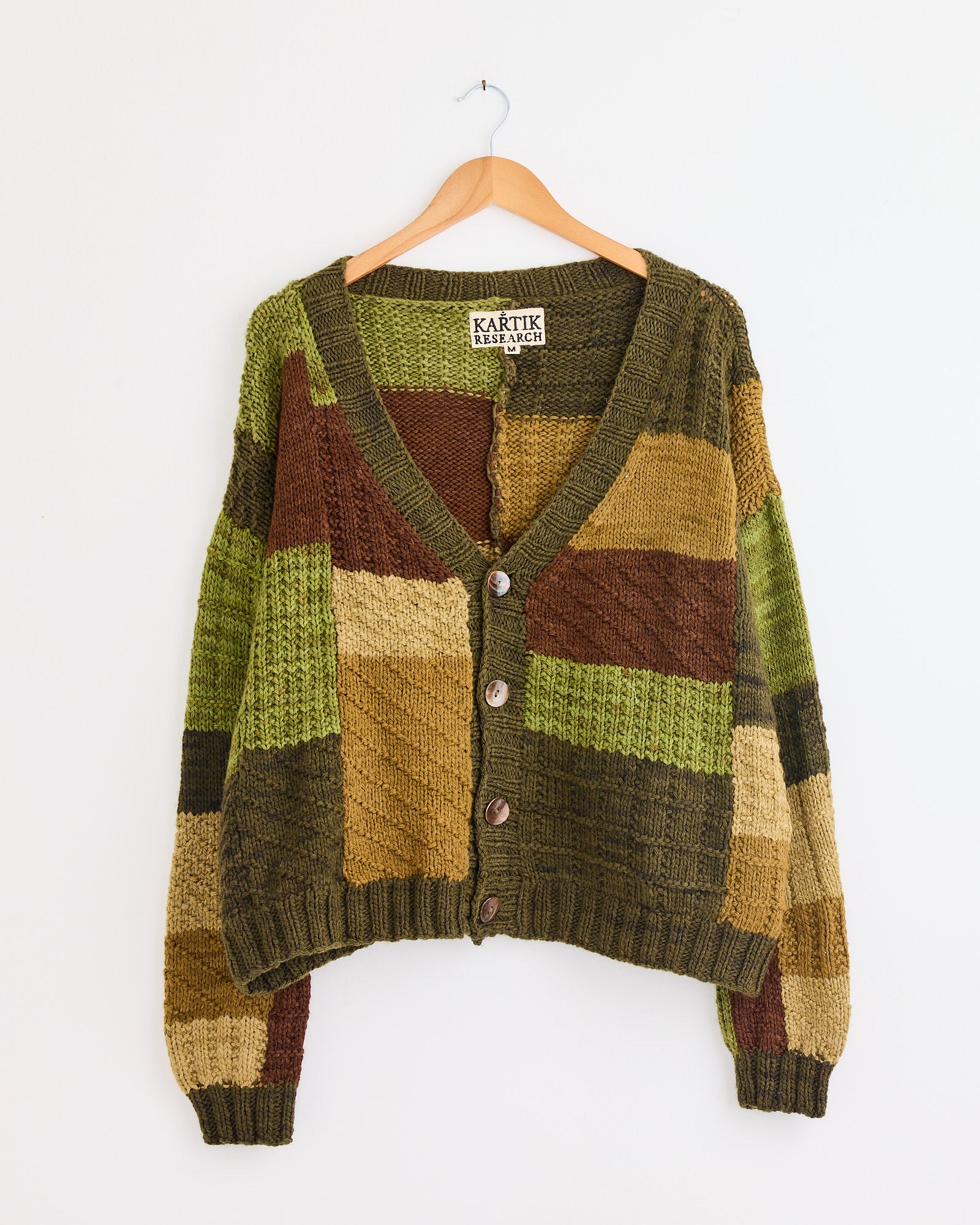 The Patchwork Cardigan in Olive/Brown/Moss by Kartik Research features hand-knit blocks of green, brown, tan, and mustard, button front, displayed on a wooden hanger against a white wall.
