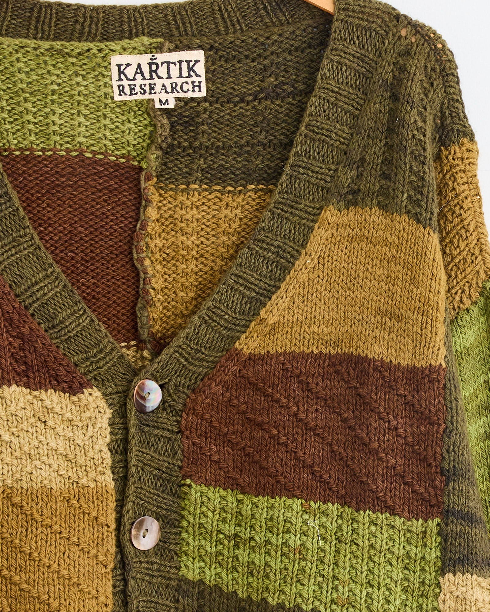 A close-up of Kartik Research’s Patchwork Cardigan in Olive/Brown/Moss, hand-knit with naturally dyed yarns. Features green, brown, mustard, and beige color blocks, two iridescent front buttons, and a Kartik Research label on the collar.