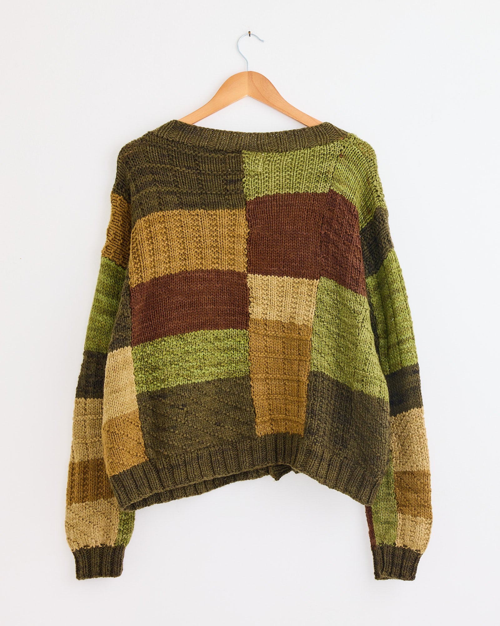 The Kartik Research Patchwork Cardigan in Olive/Brown/Moss, with a relaxed fit and long sleeves, is naturally dyed and hand-knit. It hangs on a wooden hanger against a plain white wall for cozy style.