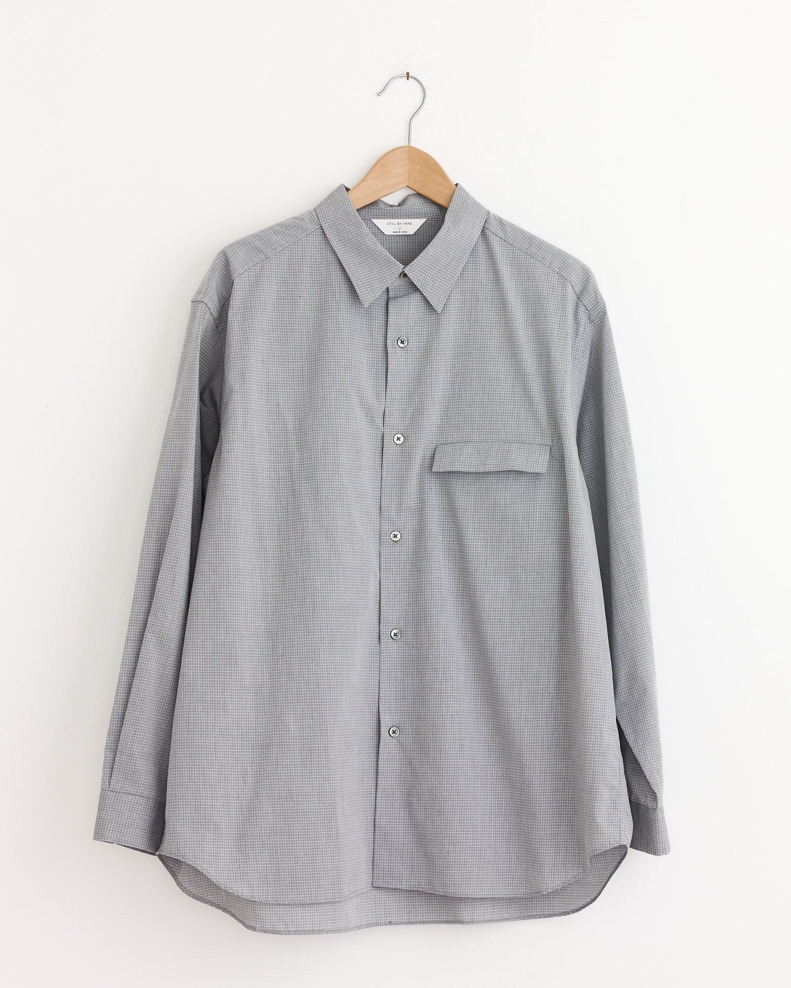 The Oversized Shirt in Grey Check by Still By Hand hangs on a wooden hanger against a plain white wall.