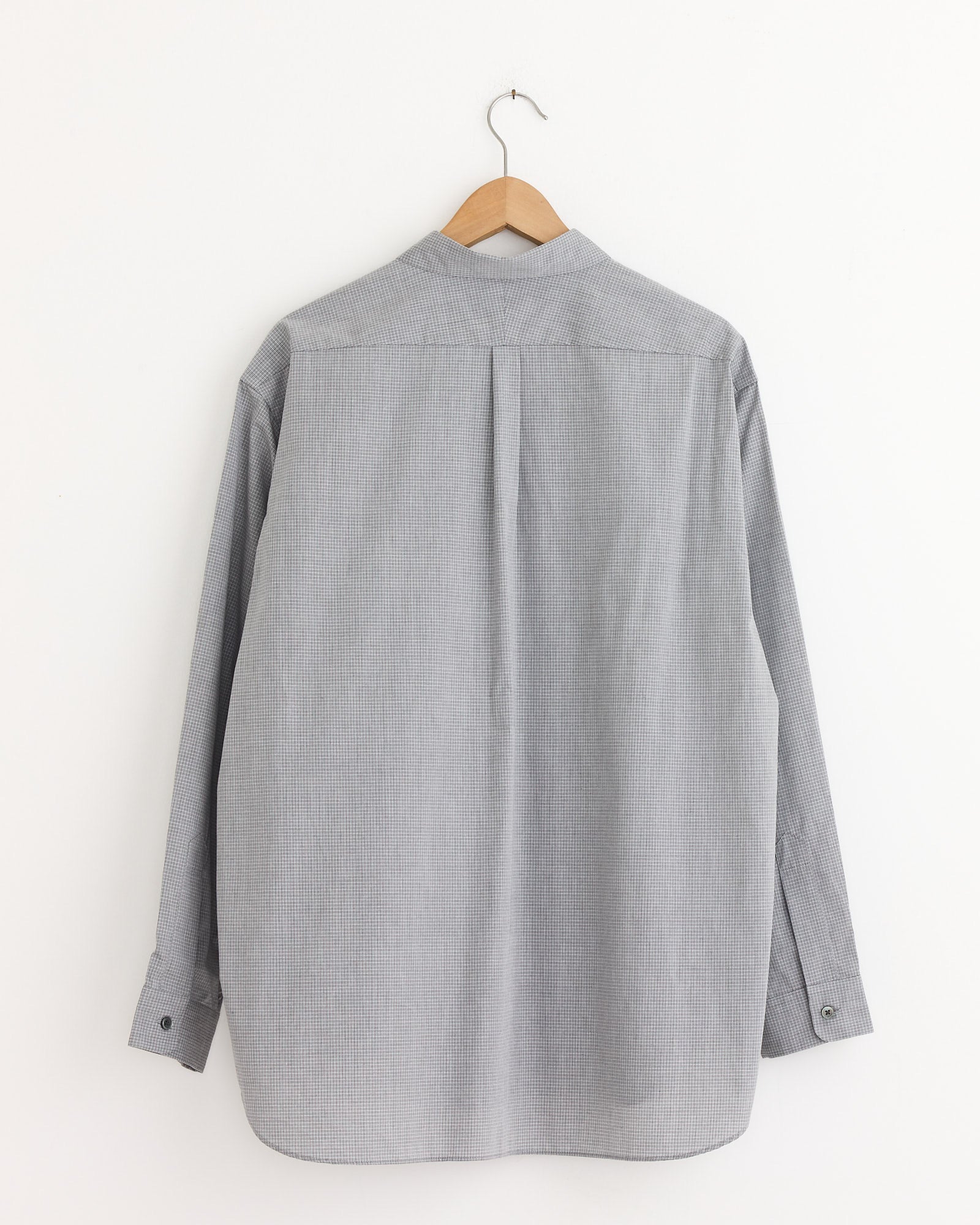 The Oversized Shirt in Grey Check by Still By Hand, featuring long sleeves and crafted in Japan, hangs on a wooden hanger against a plain white wall, displayed from the back.