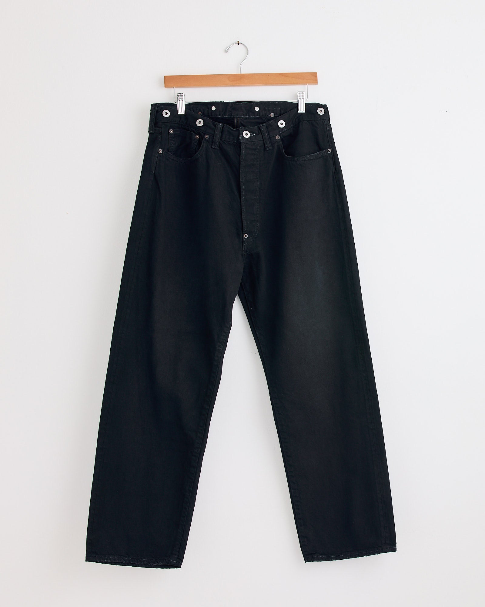 The A.PRESSE No.22 Washed Wide Denim Pant in Custom, featuring a high waist and vintage-inspired fit, hangs on a wooden hanger against a plain white wall.