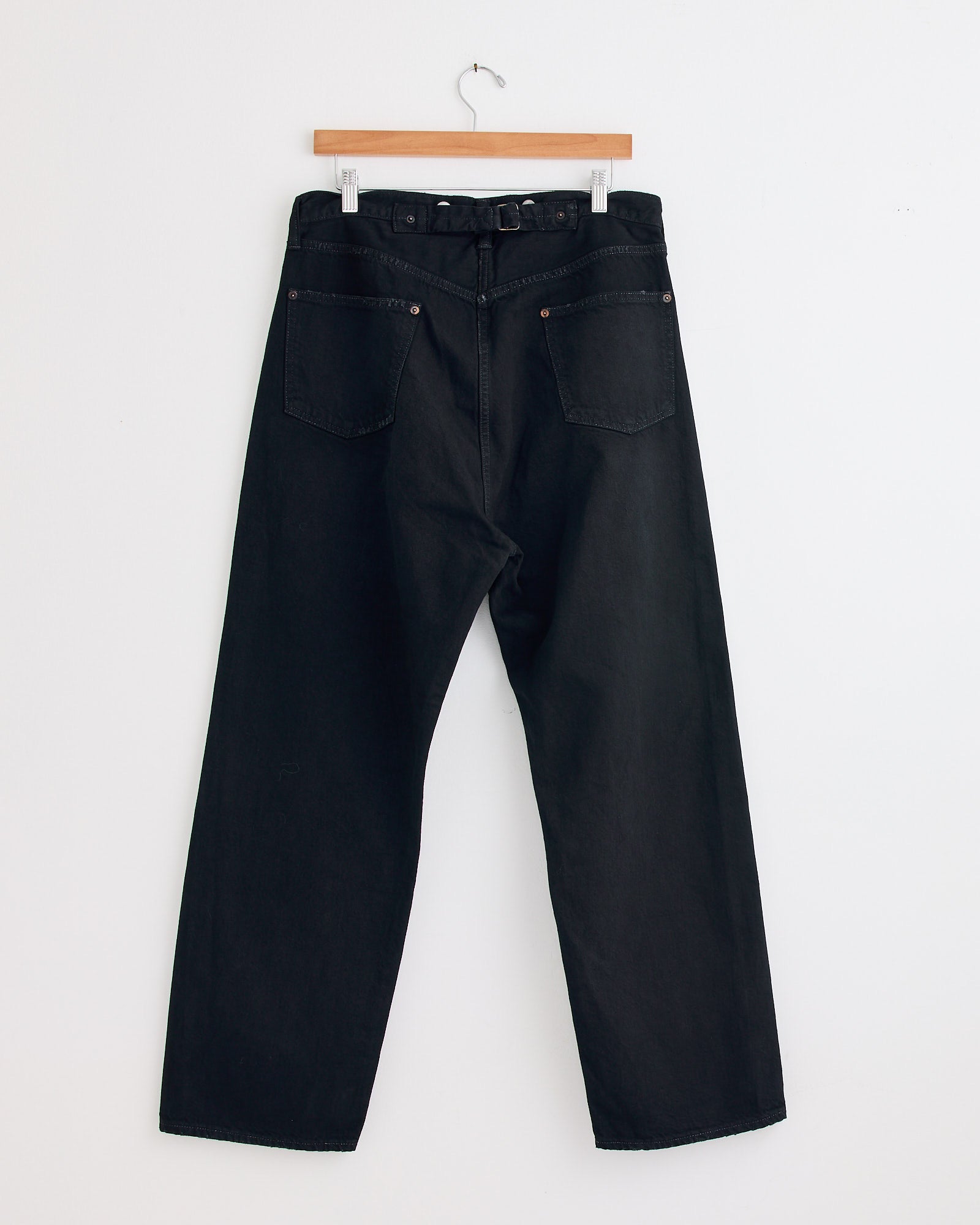 The A.PRESSE No.22 Washed Wide Denim Pant in Custom, shown with a belt on a wooden hanger against a white wall, displays the rear pockets and relaxed fit, crafted in red-selvedge indigo denim for classic style.