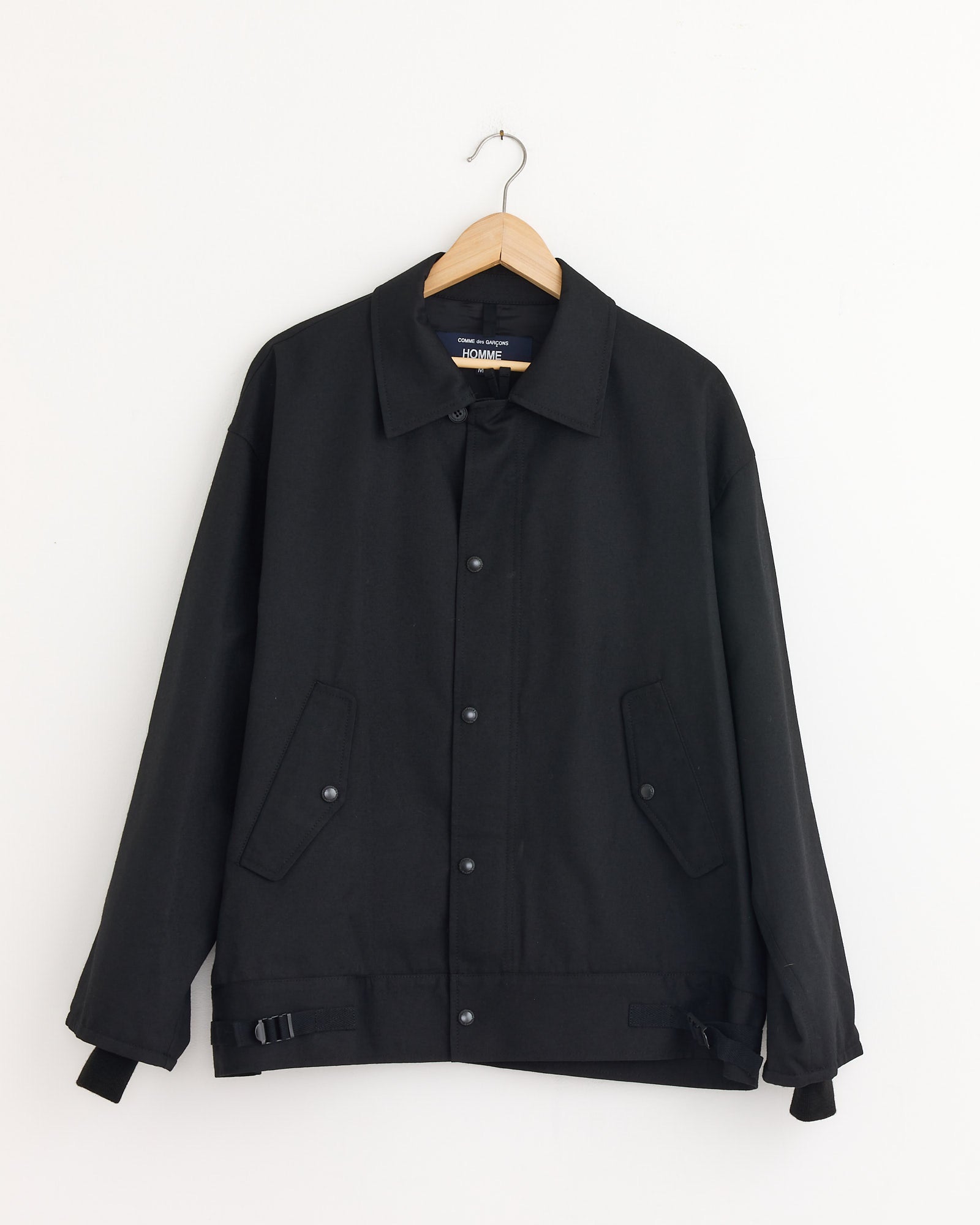 The Comme Des Garçons Homme Button Flap Pocket Jacket in Black, featuring a structured fit and buttoned front with slanted flap pockets, hangs on a wooden hanger against a plain white wall.