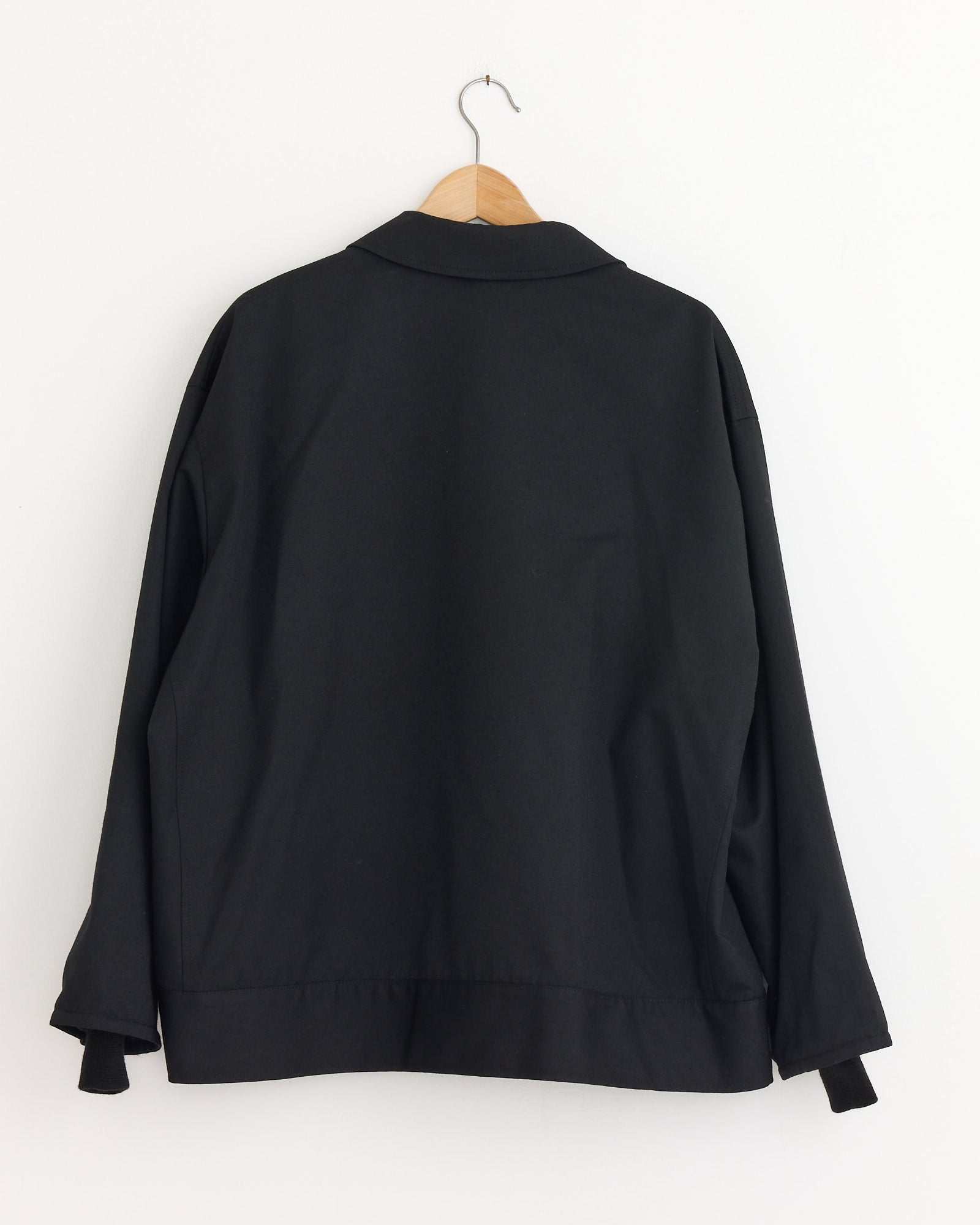 The Comme Des Garçons Homme Button Flap Pocket Jacket in Black, featuring a structured silhouette, is displayed from the back on a wooden hanger against a white wall.