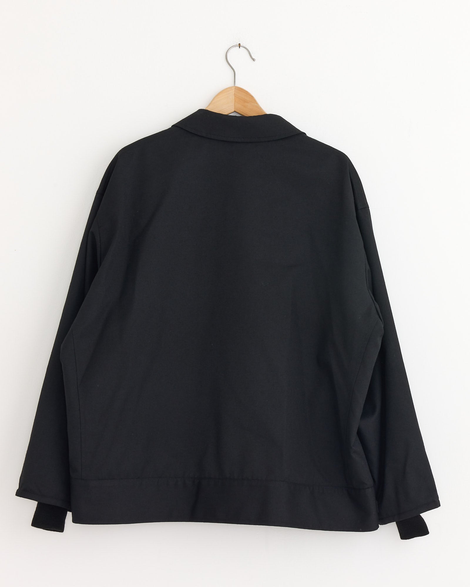 A Comme Des Garçons Homme Button Flap Pocket Jacket in black, featuring a structured silhouette, hangs on a wooden hanger and is displayed from the back against a white wall.