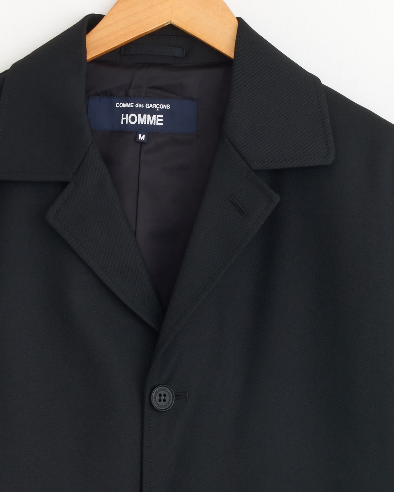 A Comme Des Garçons Homme Jacket in Black, made from 100% wool with a notched collar, hangs on a wooden hanger. The inner label displays the size: Medium.