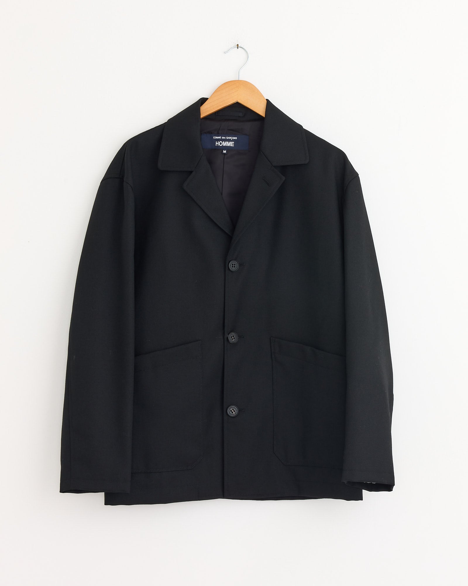 The Comme Des Garçons Homme Jacket in Black, made from 100% wool with large front pockets, hangs on a wooden hanger against a plain white wall.