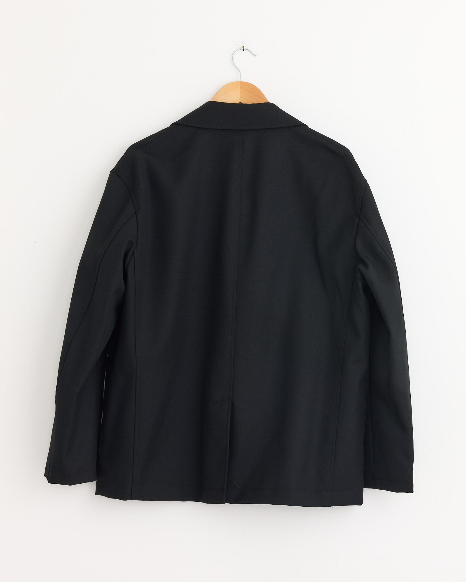 The Comme Des Garçons Homme Jacket in Black is shown from the back, featuring long sleeves, a center seam, and three buttons. Crafted from 100% wool, it hangs on a wooden hanger against a plain white wall.