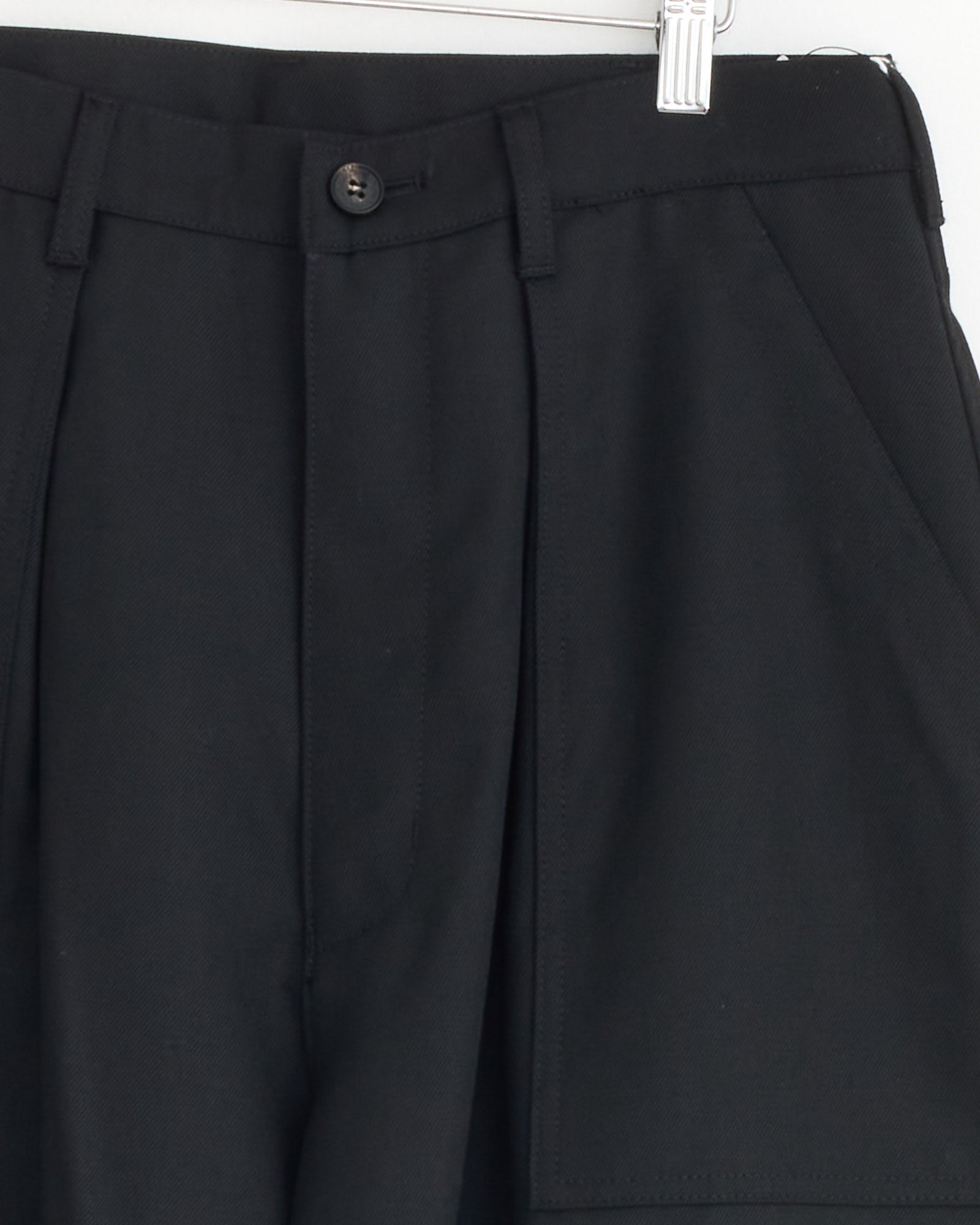Close-up of the Wool Cargo Pant in Black by Comme Des Garçons Homme, hanging on a hanger and highlighting the waistband, button, zipper area, and front pockets.
