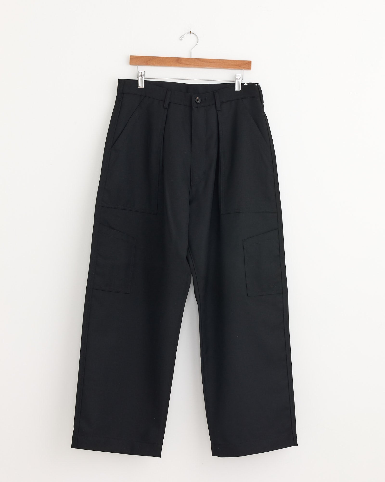 The Wool Cargo Pant in Black by Comme Des Garçons Homme, featuring front pockets, hangs on a wooden hanger against a plain white wall—a refined addition to any menswear collection.