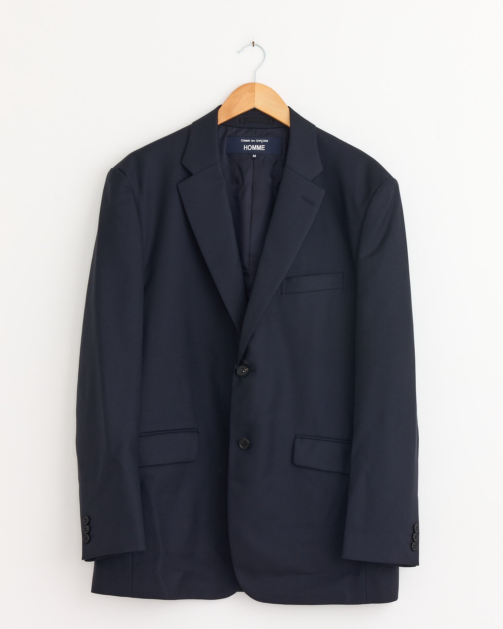 The Single-Breasted Jacket in Navy by Comme Des Garçons Homme features two buttons and flap pockets, hanging on a wooden hanger against a plain white wall.