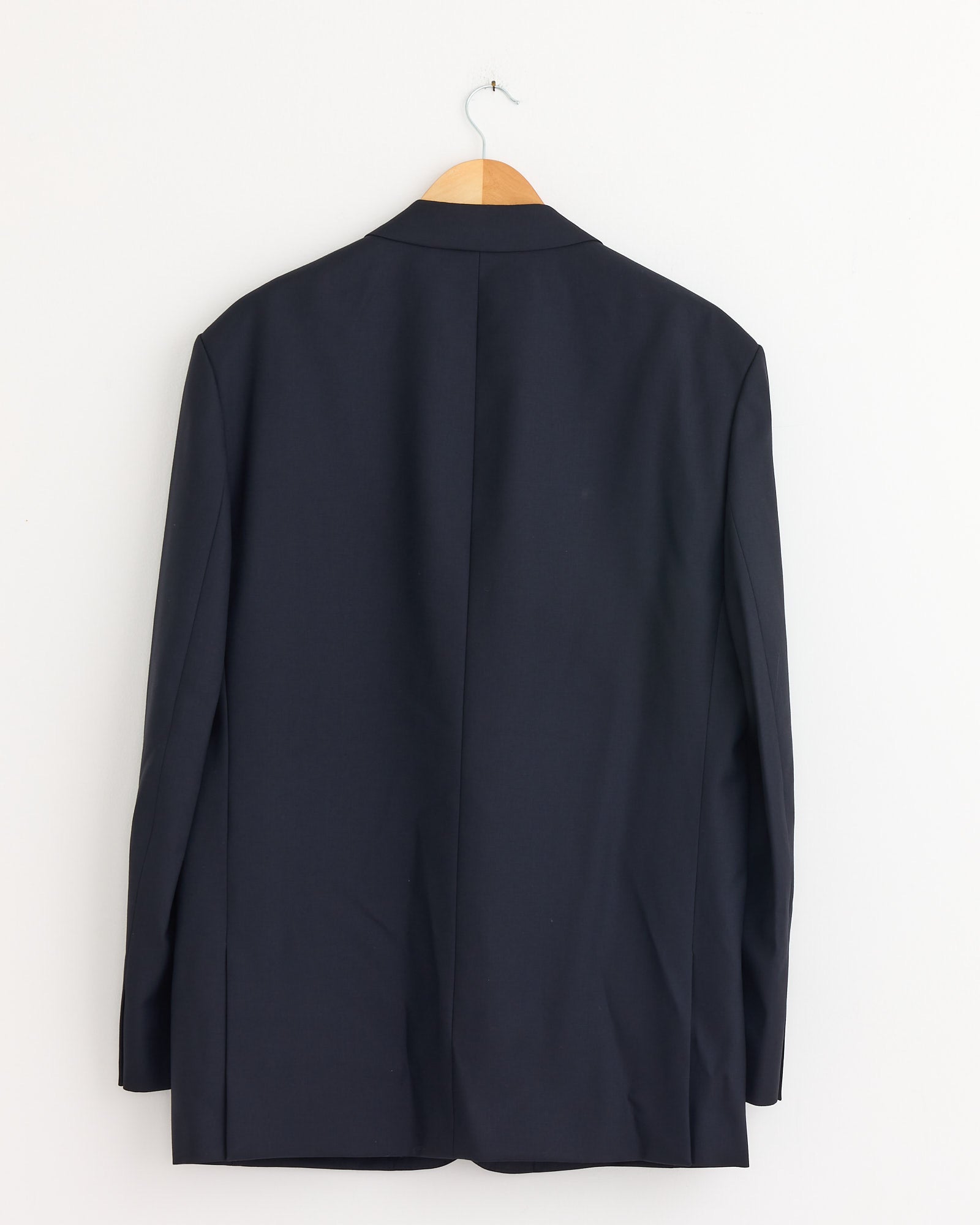 A Comme Des Garçons Homme Single-Breasted Jacket in Navy hangs from a wooden hanger against a plain white wall, displayed from the back.