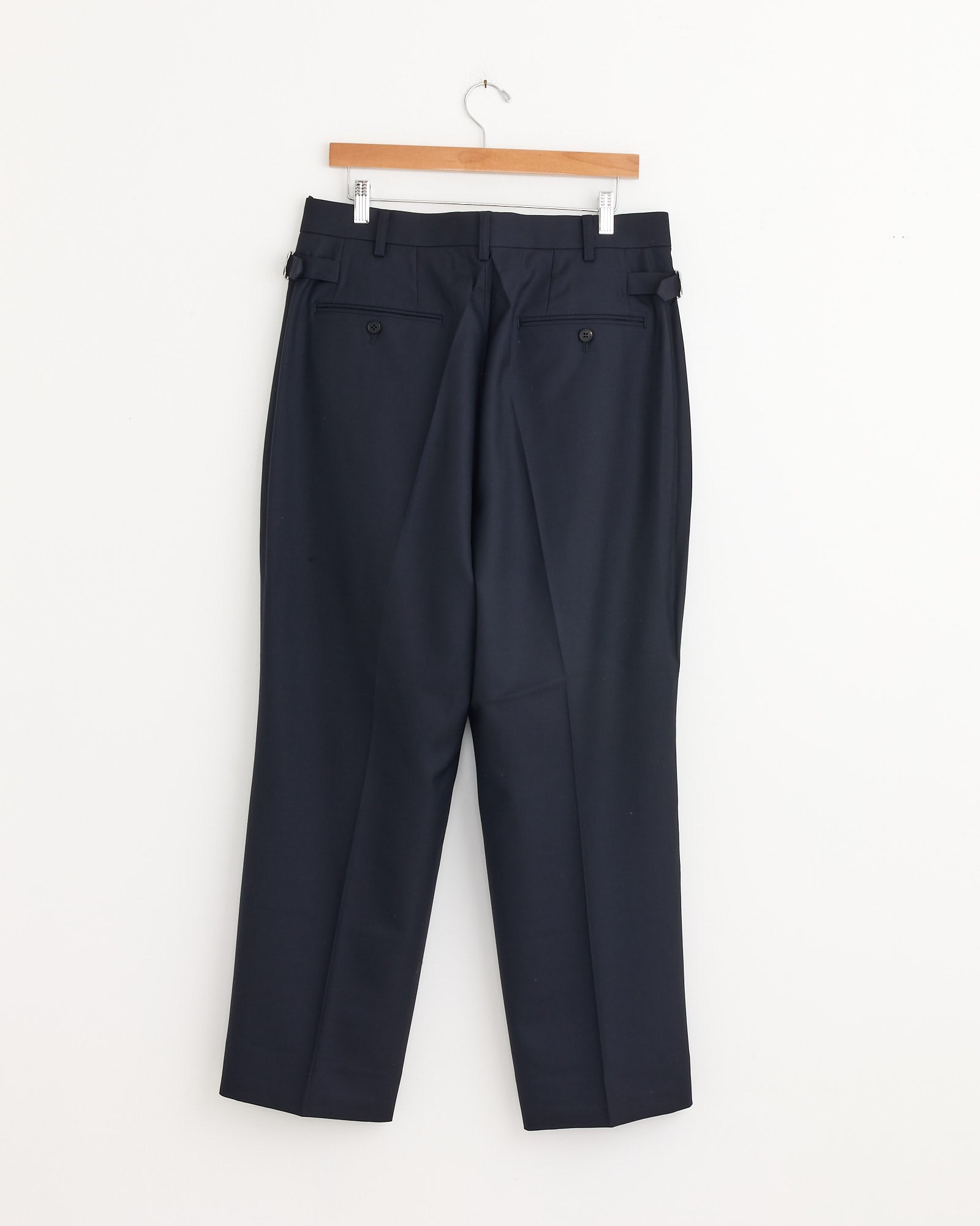 The Buckle Pant in Navy by Comme Des Garçons Homme hangs on a wooden hanger against a white wall. These tailored dress pants feature buttoned back pockets, belt loops, and offer a classic, refined style.