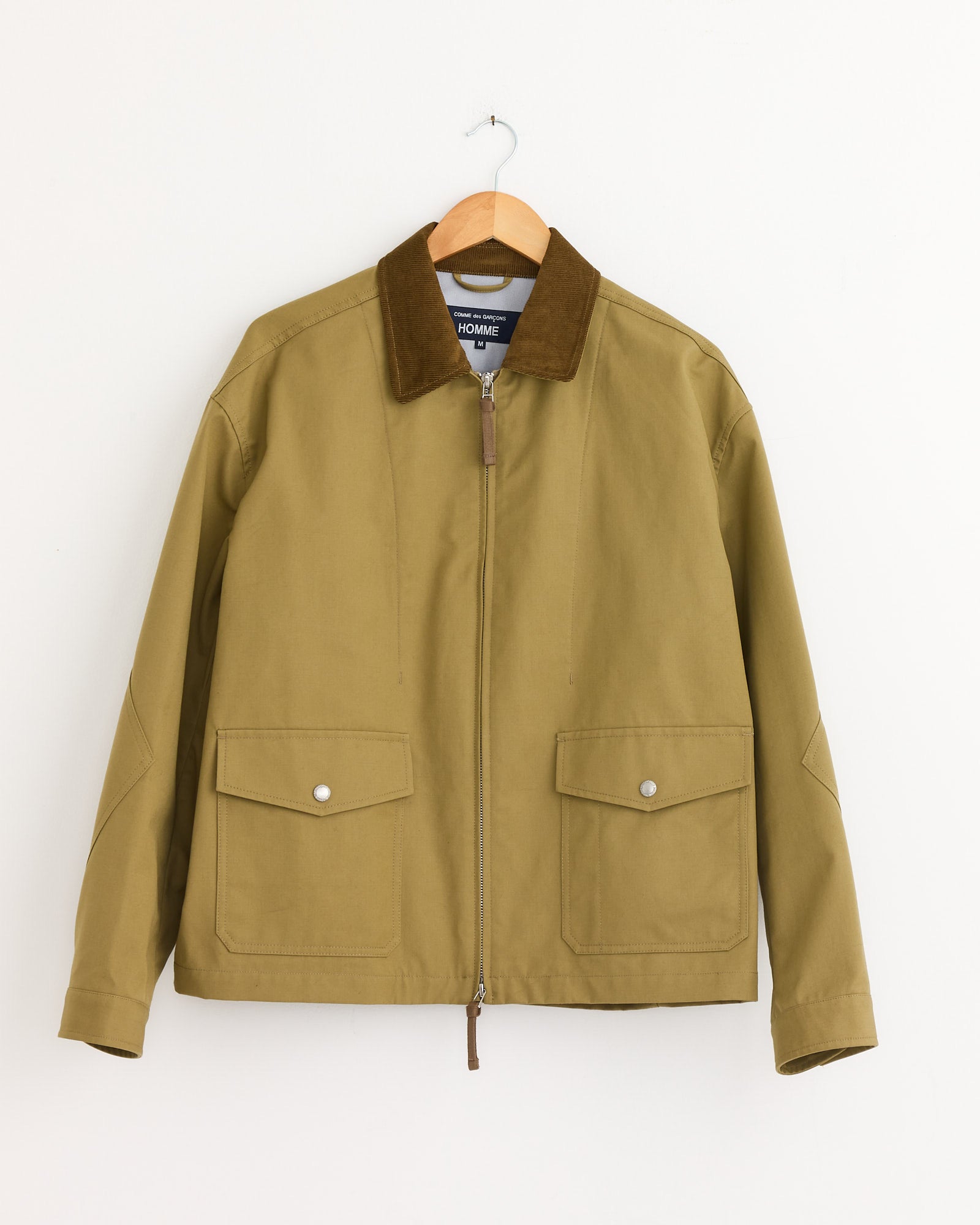 The Triple Layer Jacket in Beige by Comme Des Garçons Homme features weather-resistant fabric, a brown corduroy collar, front zipper, and two large flap pockets with silver snap buttons, displayed on a wooden hanger against a white wall.
