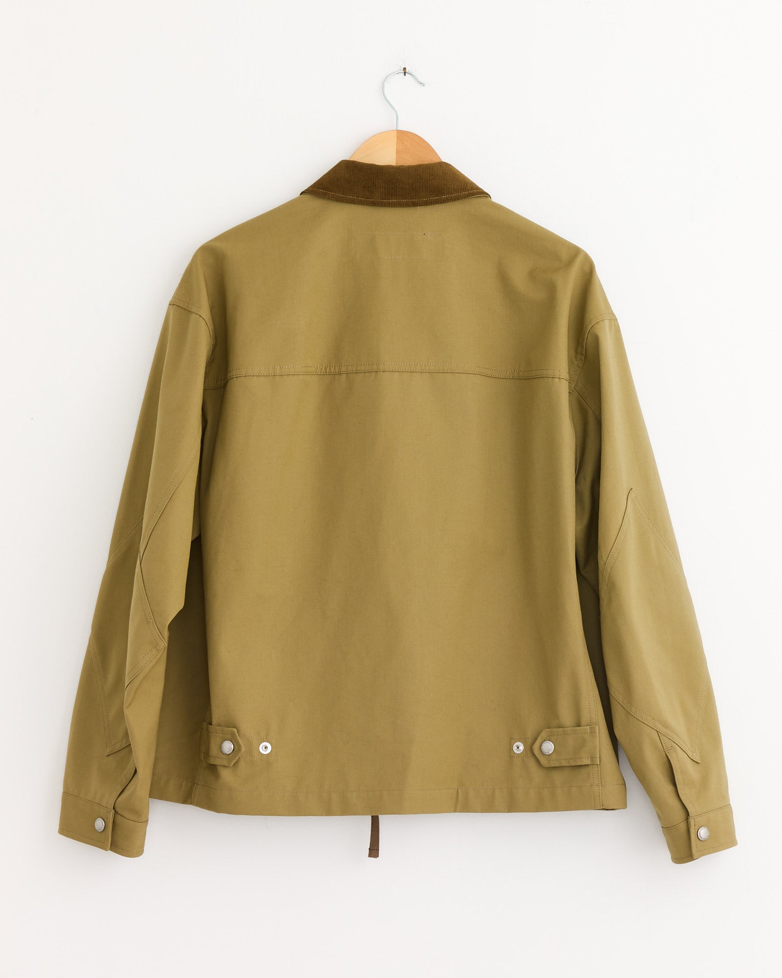 The Comme Des Garçons Homme Triple Layer Jacket in Beige hangs on a wooden hanger against a white wall, shown from the back. It features a brown collar, buttoned cuffs, and adjustable waist tabs.