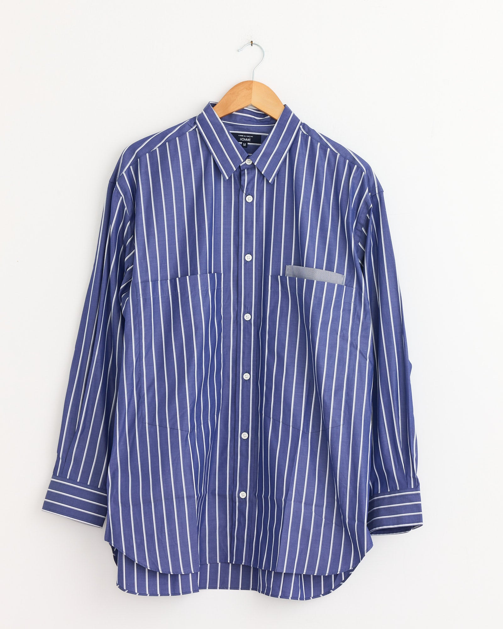 The Striped Pocket Shirt in Navy/White by Comme Des Garçons Homme hangs on a wooden hanger against a white wall. This blue, long-sleeve button-up features white vertical stripes, a collar, and a single chest pocket.