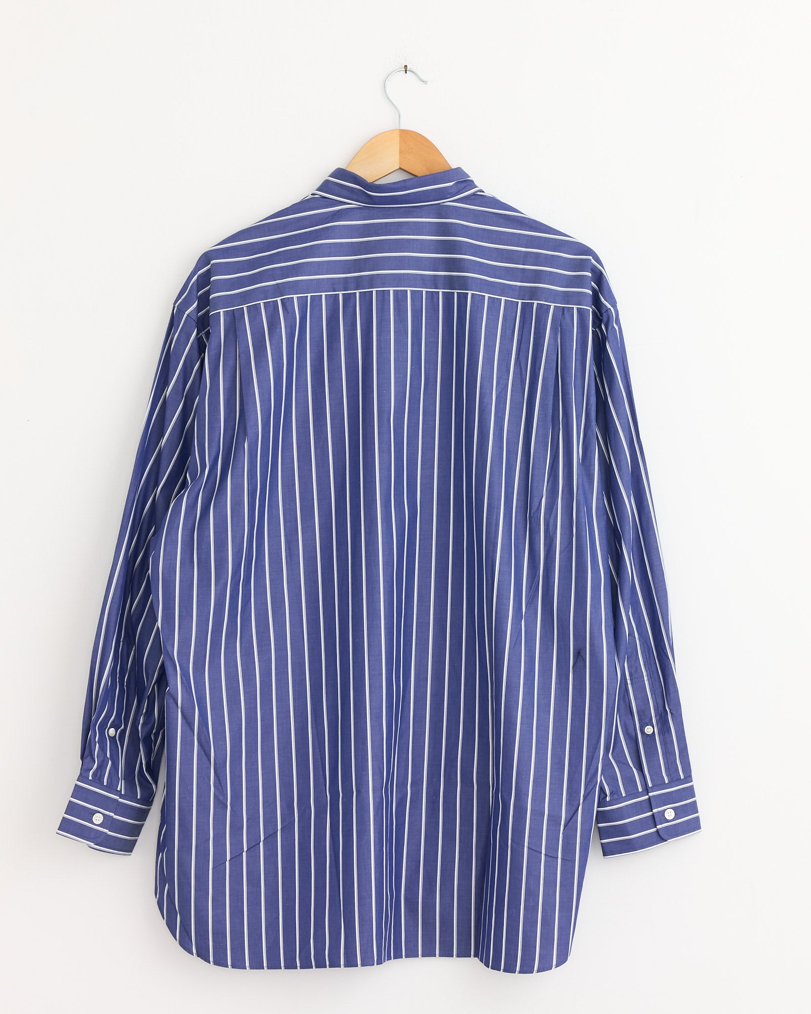 The Striped Pocket Shirt in Navy/White by Comme Des Garçons Homme hangs on a wooden hanger against a white wall, displayed from the back to show its long sleeves, buttoned cuffs, and crisp vertical stripes.
