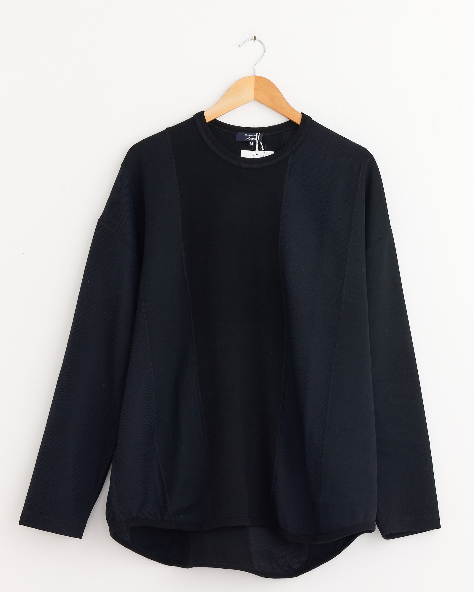 The Panelled T-Shirt in Black Mix by Comme Des Garçons Homme hangs on a wooden hanger against a white wall. Made in Japan, it features a relaxed fit and a slightly curved hem.