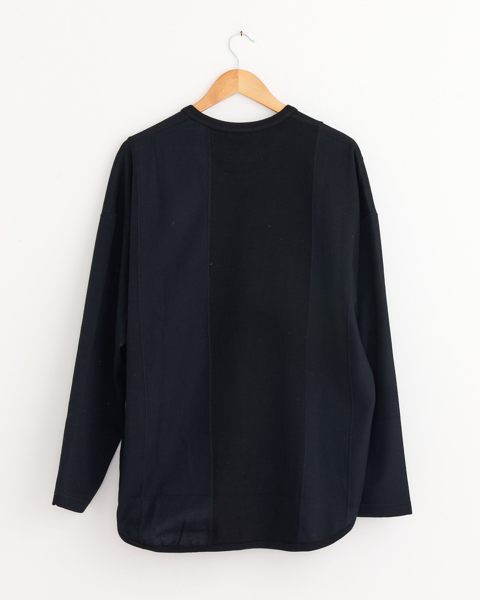 The Panelled T-Shirt in Black Mix by Comme Des Garçons Homme hangs on a wooden hanger against a plain white wall, shown from the back.
