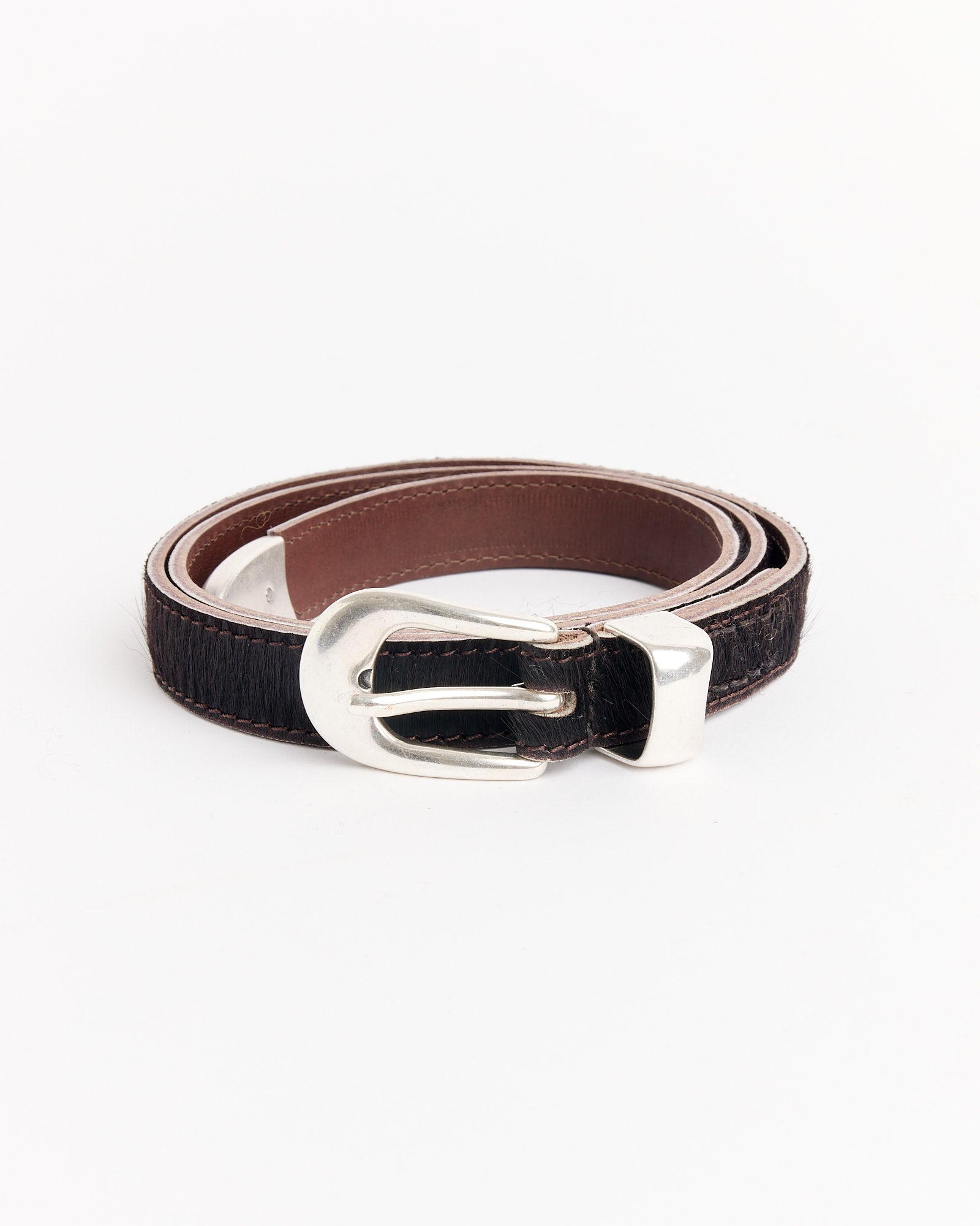 The Our Legacy 2 CM Belt in Chocolate, featuring a shiny silver buckle, is shown coiled on a white background.