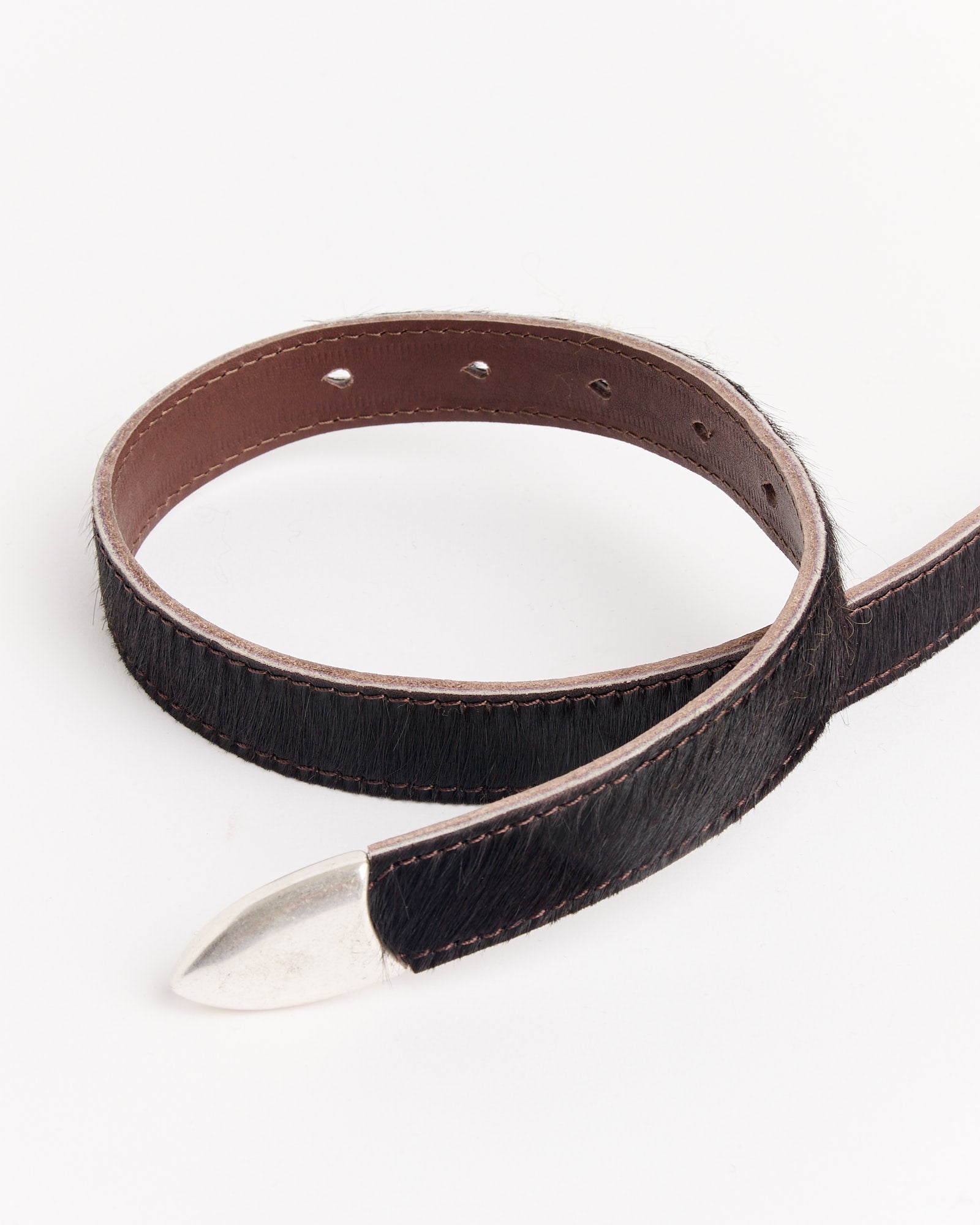 The Our Legacy 2 CM Belt in Chocolate features a coiled black leather design with a brown inner lining and a shiny silver pointed tip, displayed on a white background.