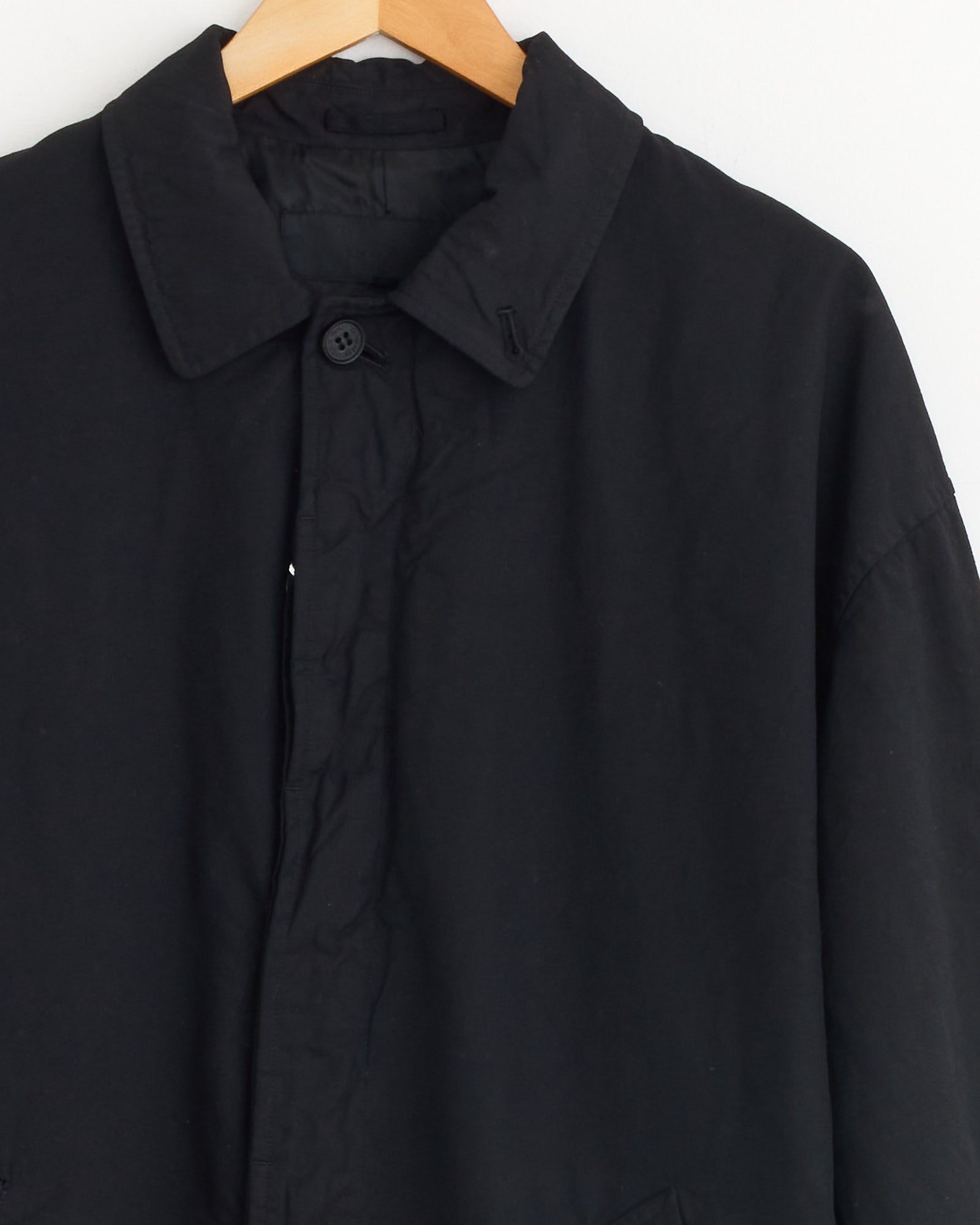 The Comme Des Garçons Homme Mac Coat in Black hangs buttoned on a wooden hanger against a white background, showcasing its simple, classic design.
