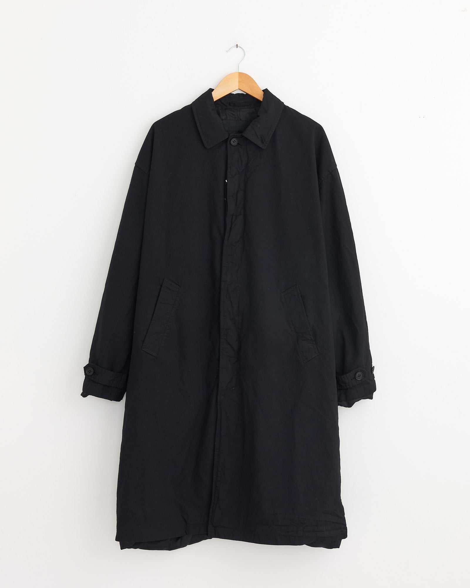 The Mac Coat in Black by Comme Des Garçons Homme hangs on a wooden hanger against a white wall, featuring a collar, buttoned front, two pockets, and refined buttoned cuffs for a classic look.