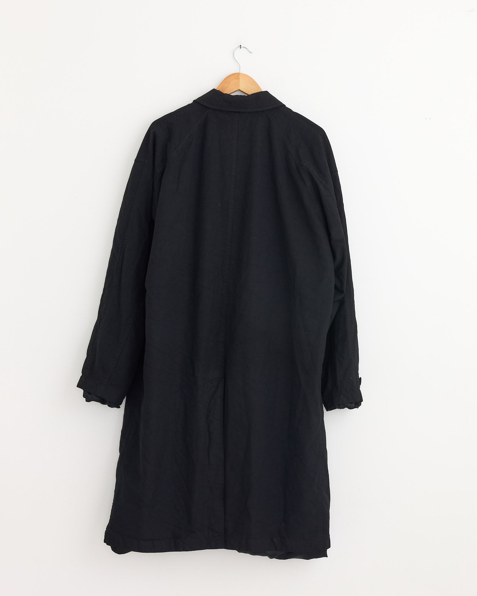 The Mac Coat in Black by Comme Des Garçons Homme, featuring long sleeves, is displayed on a wooden hanger against a plain white wall with its back facing forward.