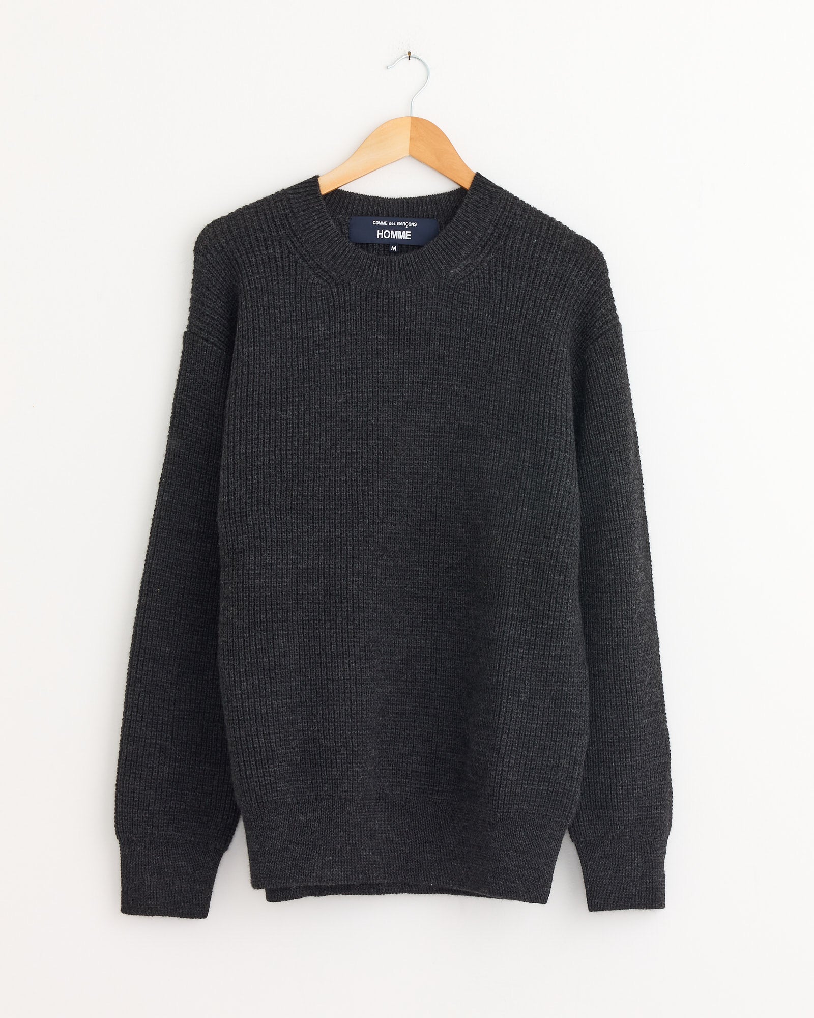 The Knit Sweater in Top Grey by Comme Des Garçons Homme, a heavyweight wool knit in dark grey, hangs on a wooden hanger against a plain white wall.