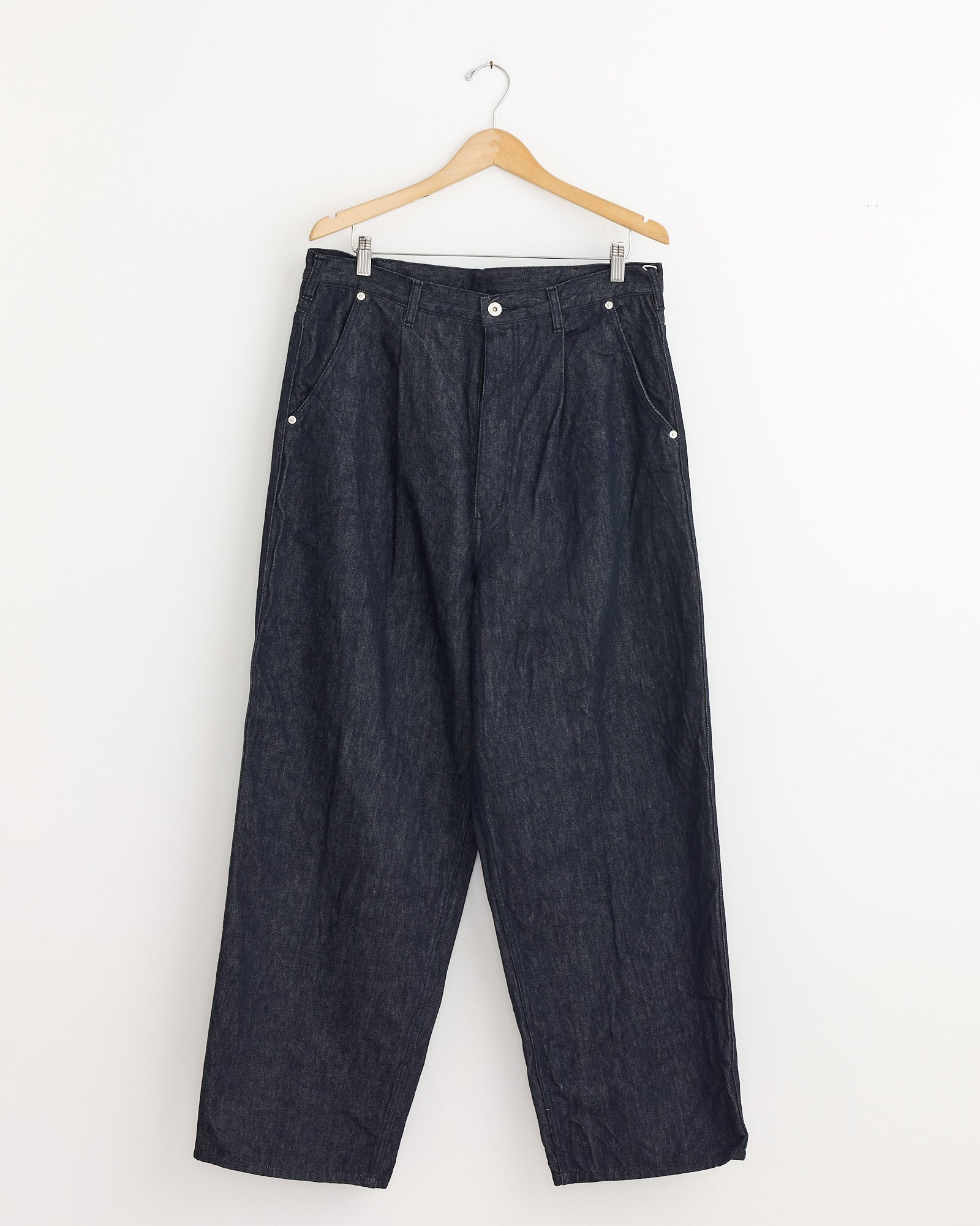 The Pleated Jeans in Indigo by Comme Des Garçons Homme, featuring a wide-leg silhouette, are made in Japan and hang on a wooden hanger against a plain white wall.
