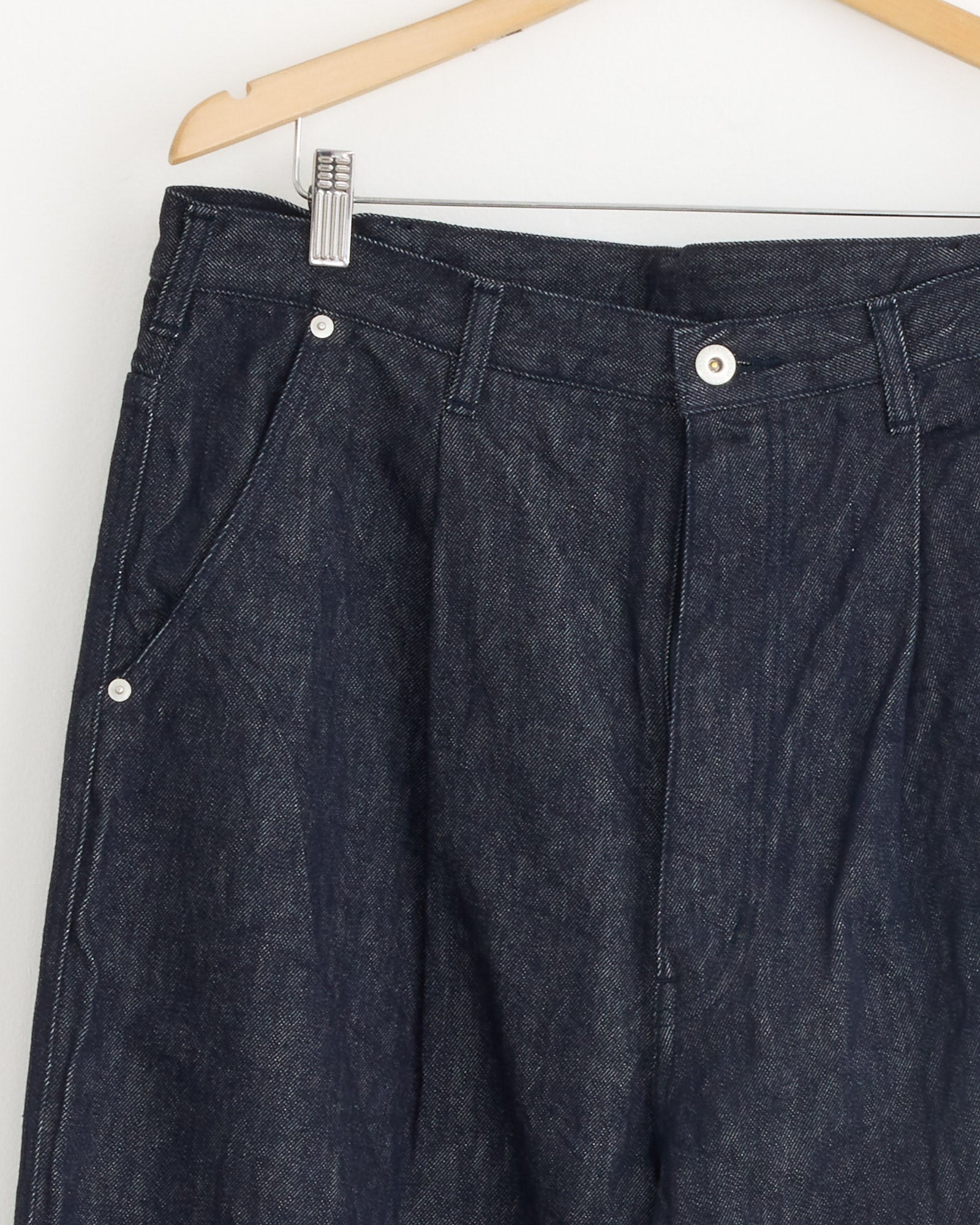 Comme Des Garçons Homme Pleated Jeans in Indigo hang on a wooden hanger against a white wall, displaying the upper half with visible front pockets, belt loops, and button closure.