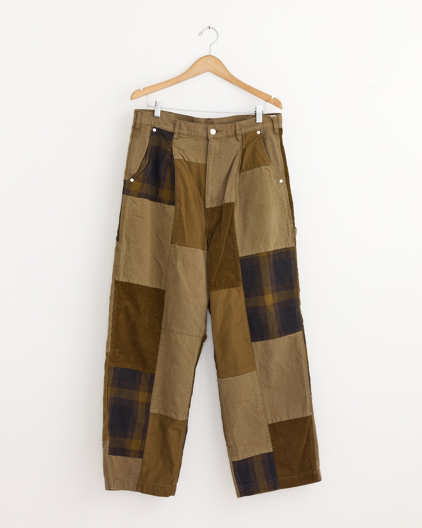 The Patchwork Pant in Beige Mix by Comme Des Garçons Homme, featuring plaid and solid panels in brown, olive, and tan tones, hangs on a wooden hanger against a plain white background.