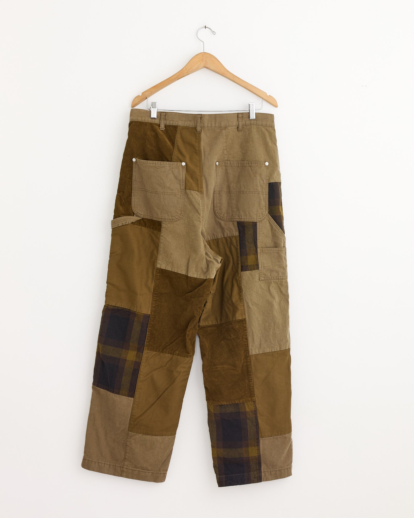 The Comme Des Garçons Homme Patchwork Pant in Beige Mix features workwear-inspired tailoring with brown, tan, olive, and plaid fabrics, two large back pockets, and a relaxed fit. Shown on a wooden hanger against a white wall.