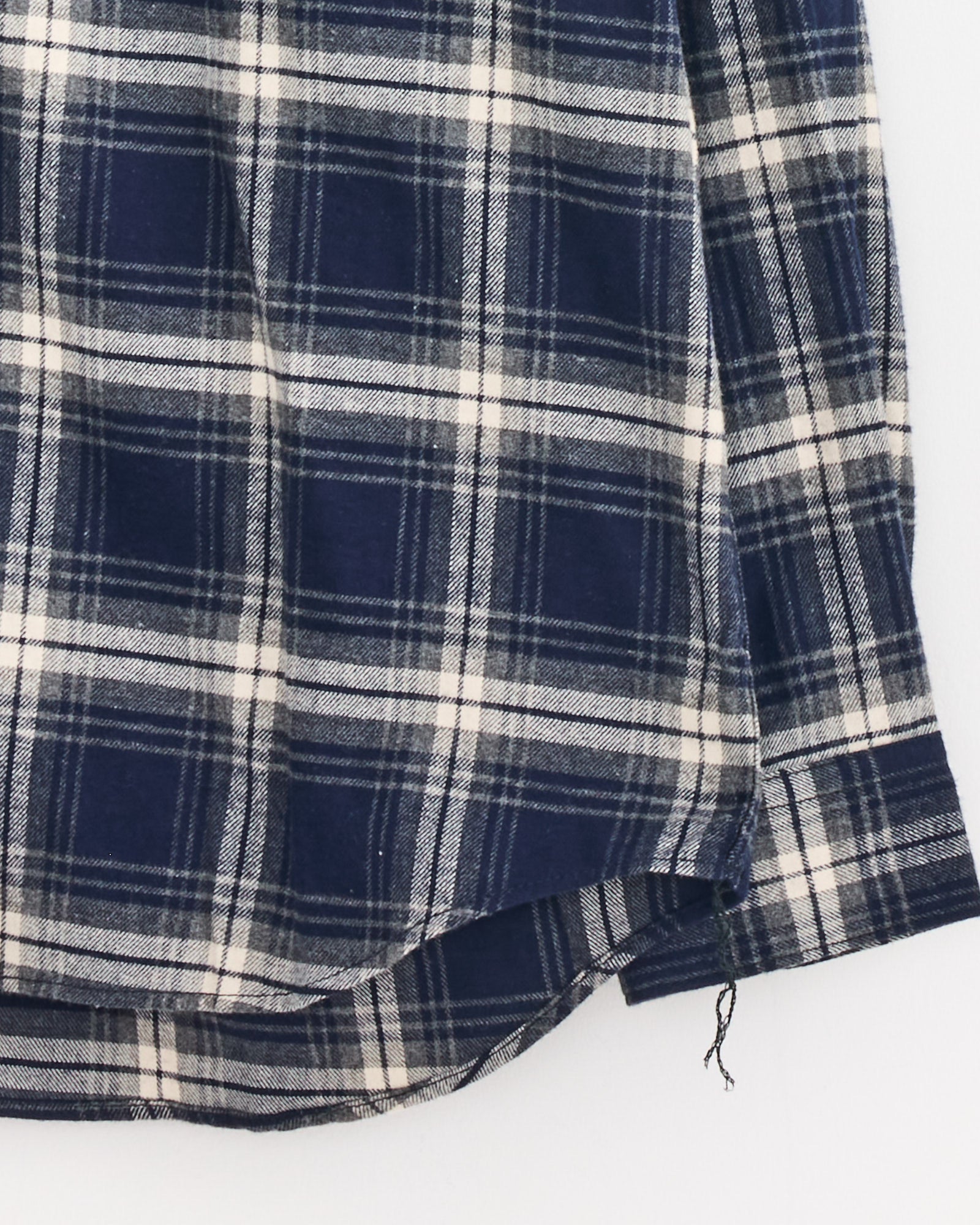 A close-up of the Double Check Shirt in Navy/Grey by Comme Des Garçons Homme, showcasing its cotton navy and grey check pattern, rounded hem, and a loose thread hanging from the bottom edge.