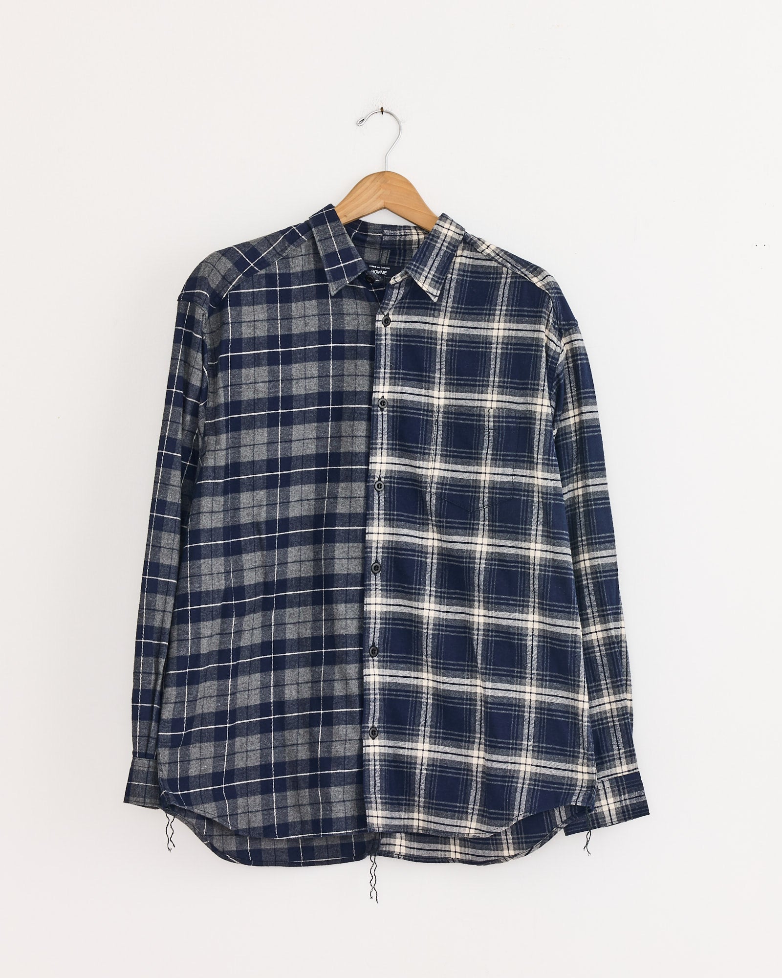A Comme Des Garçons Homme Double Check Shirt in Navy/Grey hangs on a wooden hanger, its long sleeves and split blue plaid design in quality cotton displayed against a plain white background.