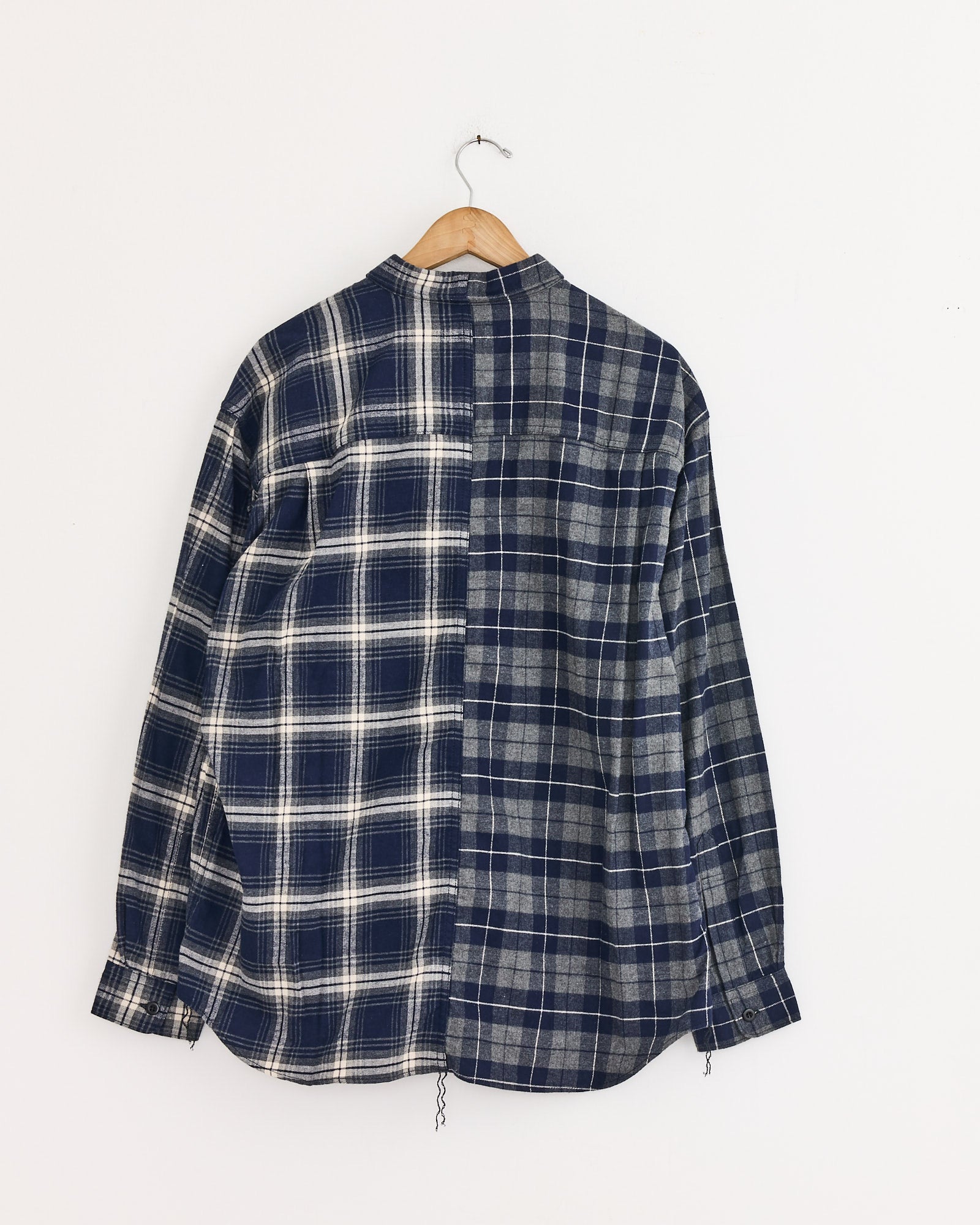 The Comme Des Garçons Homme Double Check Shirt in Navy/Grey, a cotton long-sleeve with two contrasting blue plaid patterns split vertically, hangs on a wooden hanger against a plain white wall.