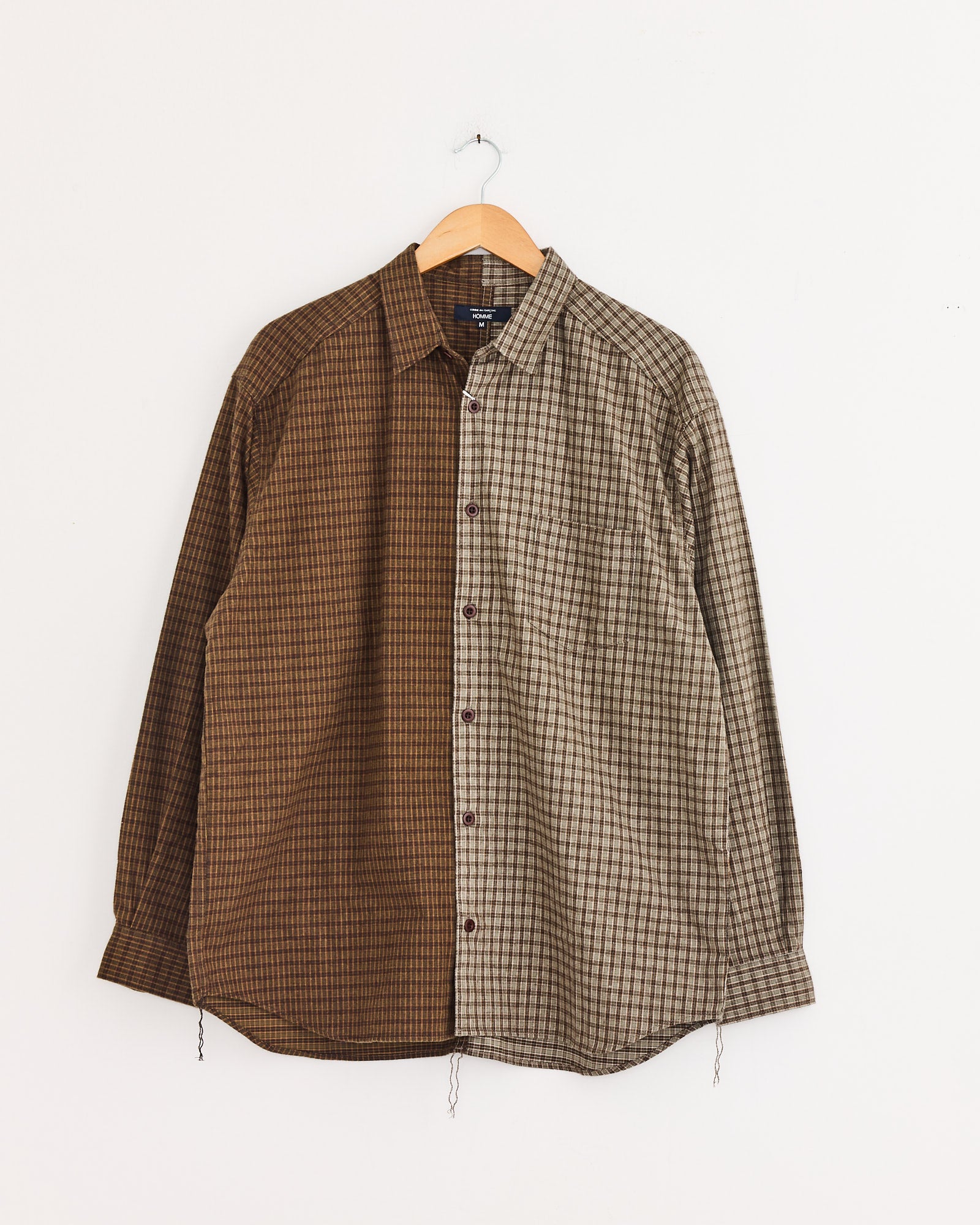 The Double Check Shirt in Khaki/Brown by Comme Des Garçons Homme is a cotton, long-sleeve shirt with a vertically split small check pattern in brown and beige, displayed on a wooden hanger against a white wall.