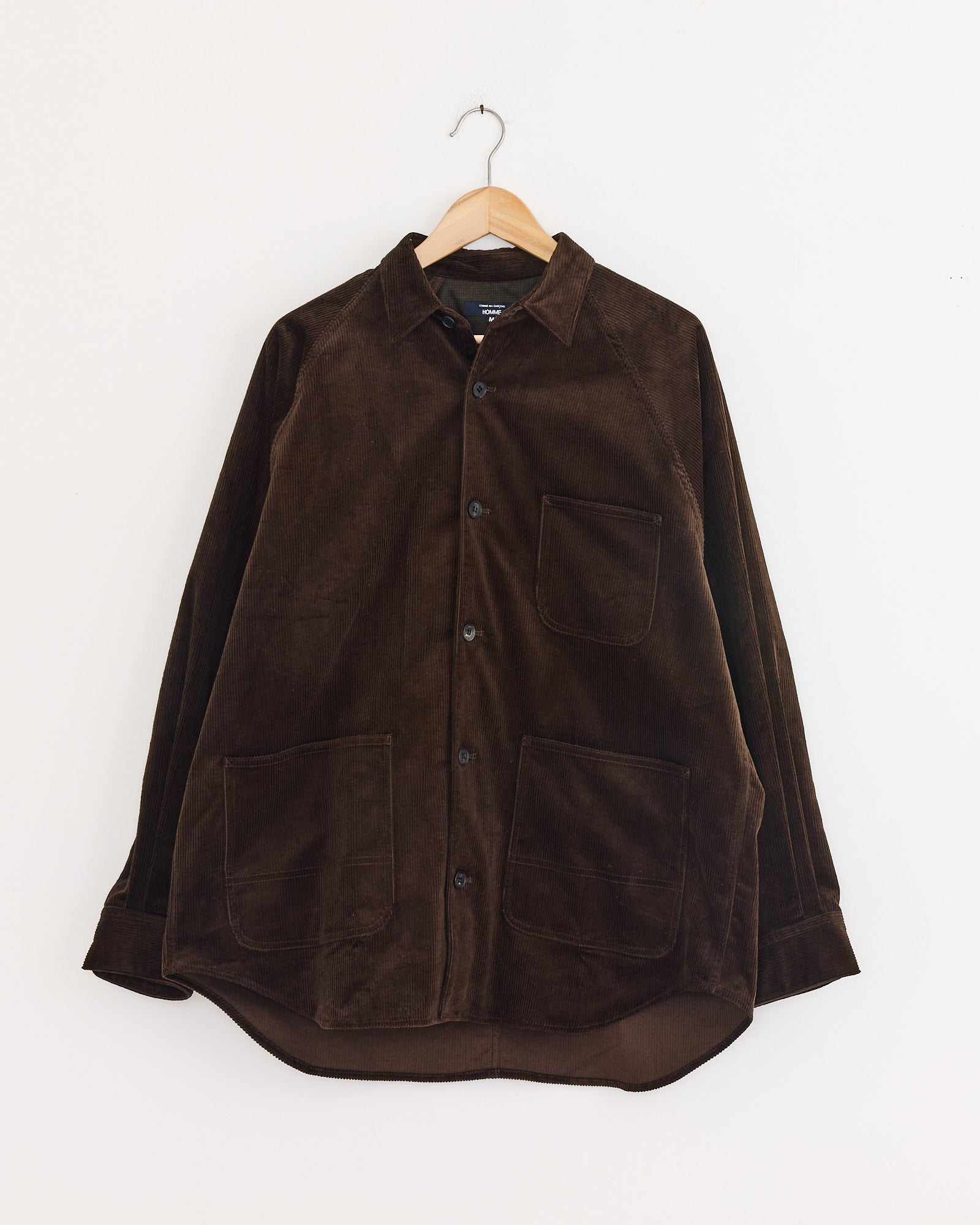 The Comme Des Garçons Homme Needlecord Shirt in Brown features long sleeves, three front patch pockets, and black buttons. Displayed on a wooden hanger against a white background. Made in Japan.