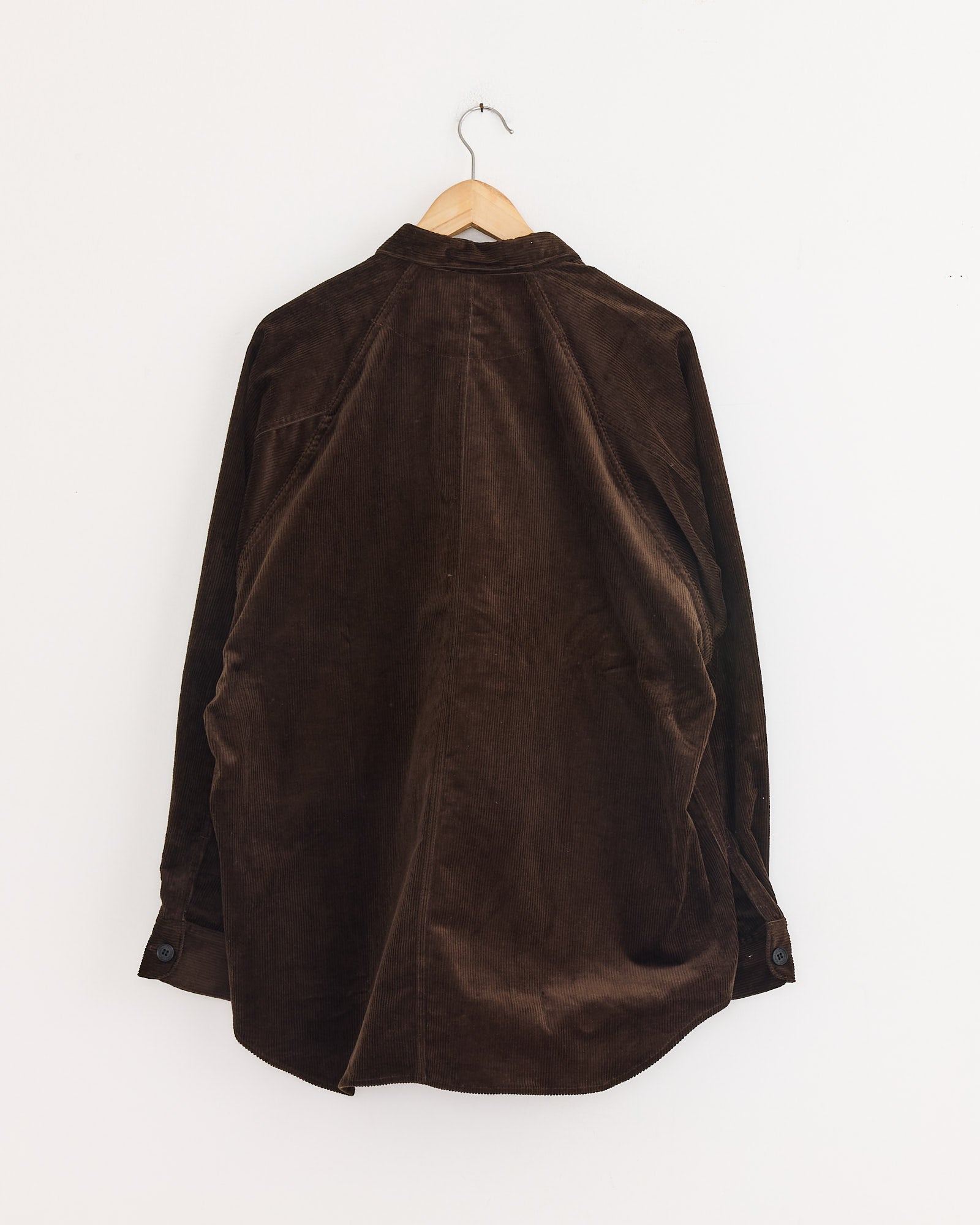 A Comme Des Garçons Homme Needlecord Shirt in brown hangs on a wooden hanger against a plain white wall, displayed from the back.