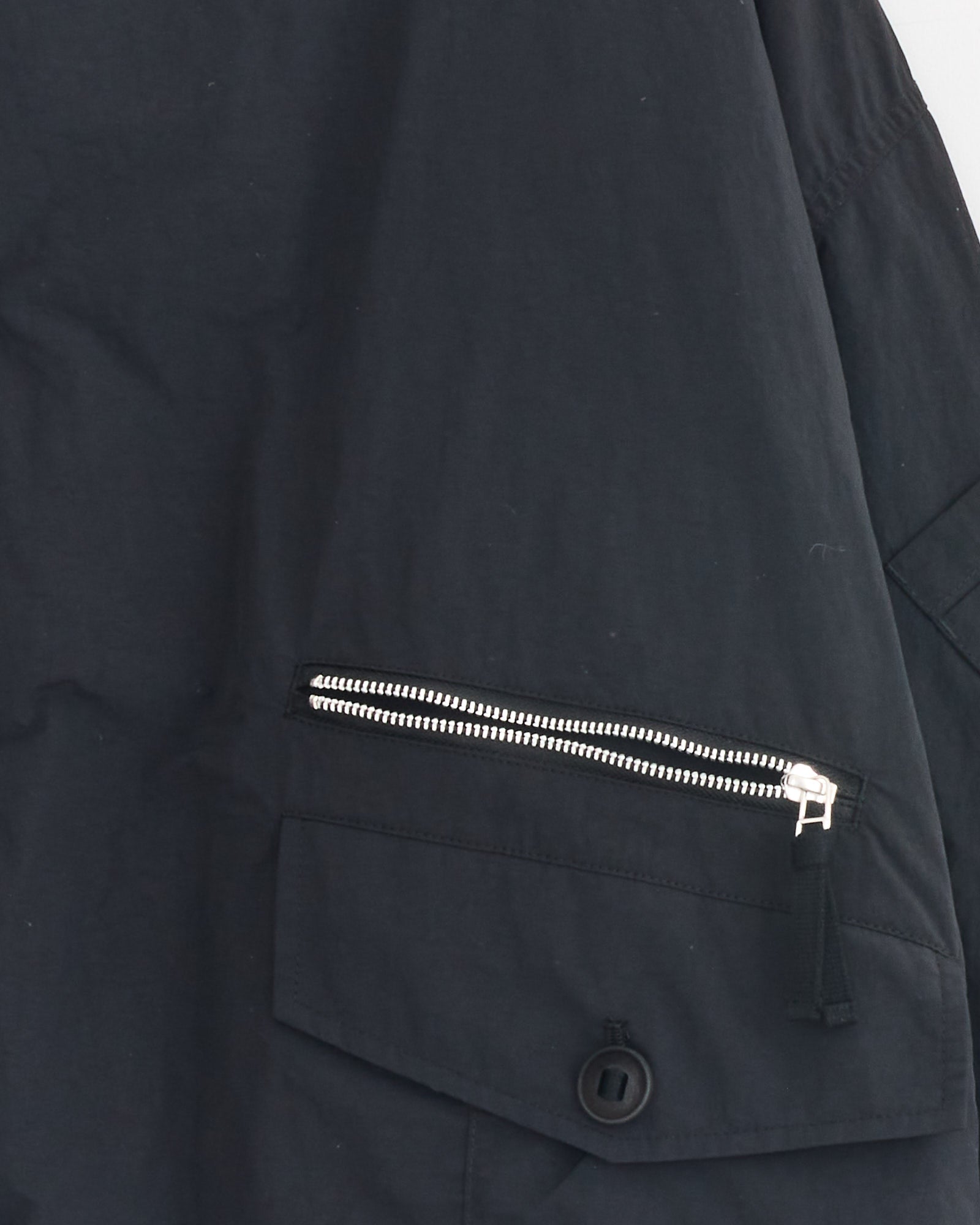 Close-up of the Comme Des Garçons Homme Utility Jacket in Black, made from nylon with a visible texture. Features include a silver zipper pocket and a flap pocket with black button below.