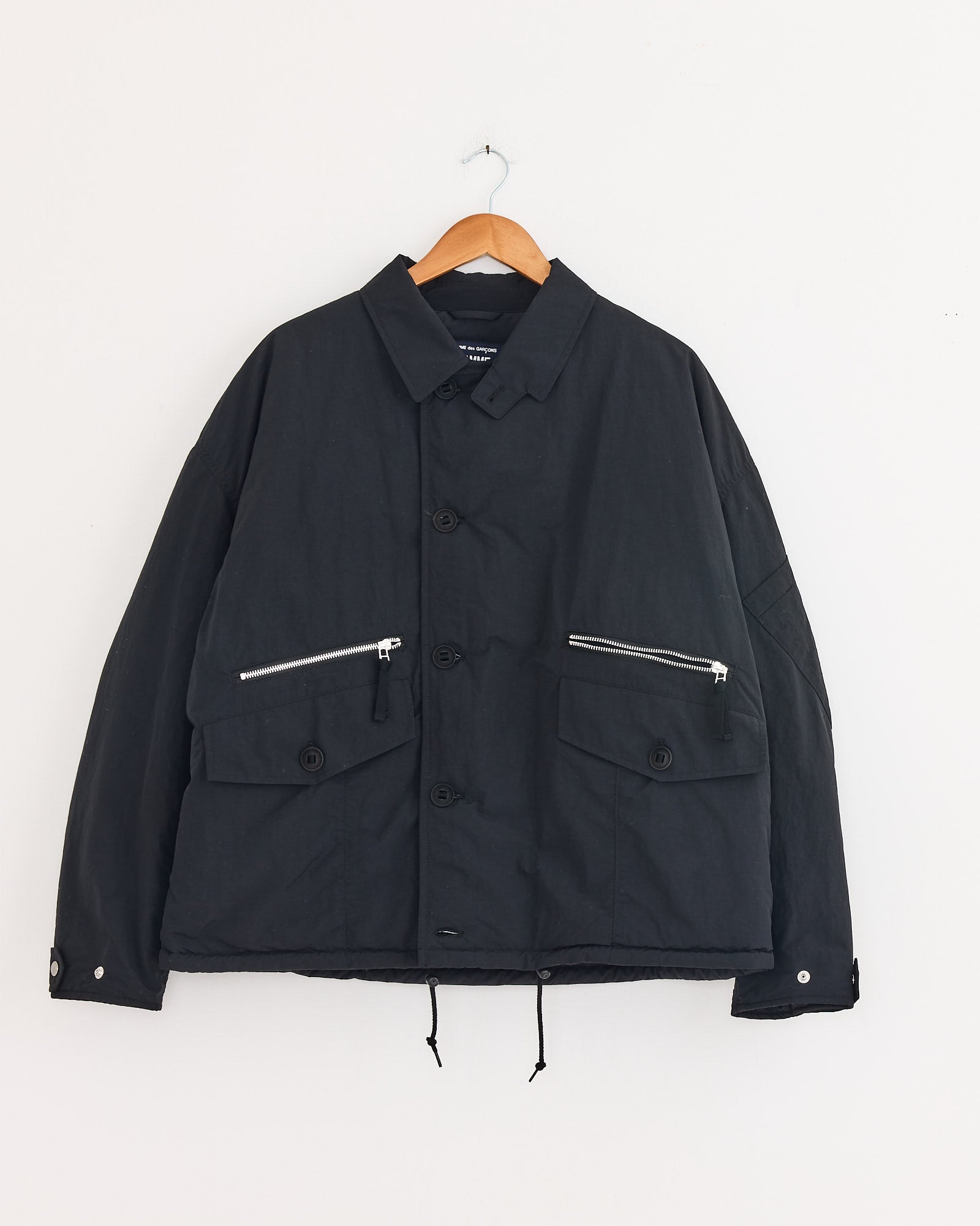 The Comme Des Garçons Homme Utility Jacket in Black, made of nylon with a collar, large button-flap front pockets, zip chest pockets, and hem drawstrings, is displayed on a wooden hanger against a white wall.