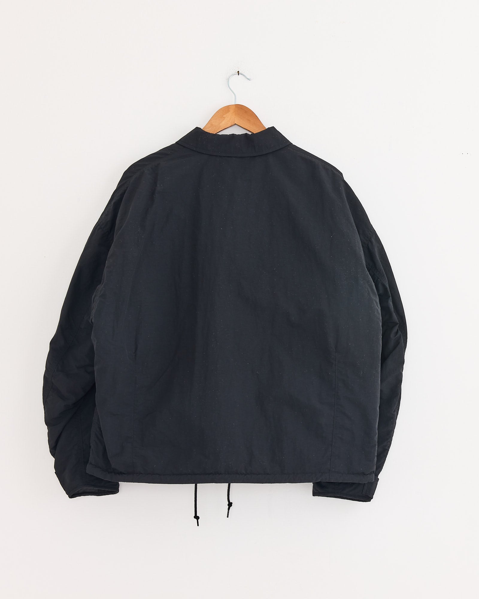 The Comme Des Garçons Homme Utility Jacket in Black, made of nylon, hangs on a wooden hanger against a white wall. Shown from the back, it features a collar, long sleeves, and a drawstring hem.