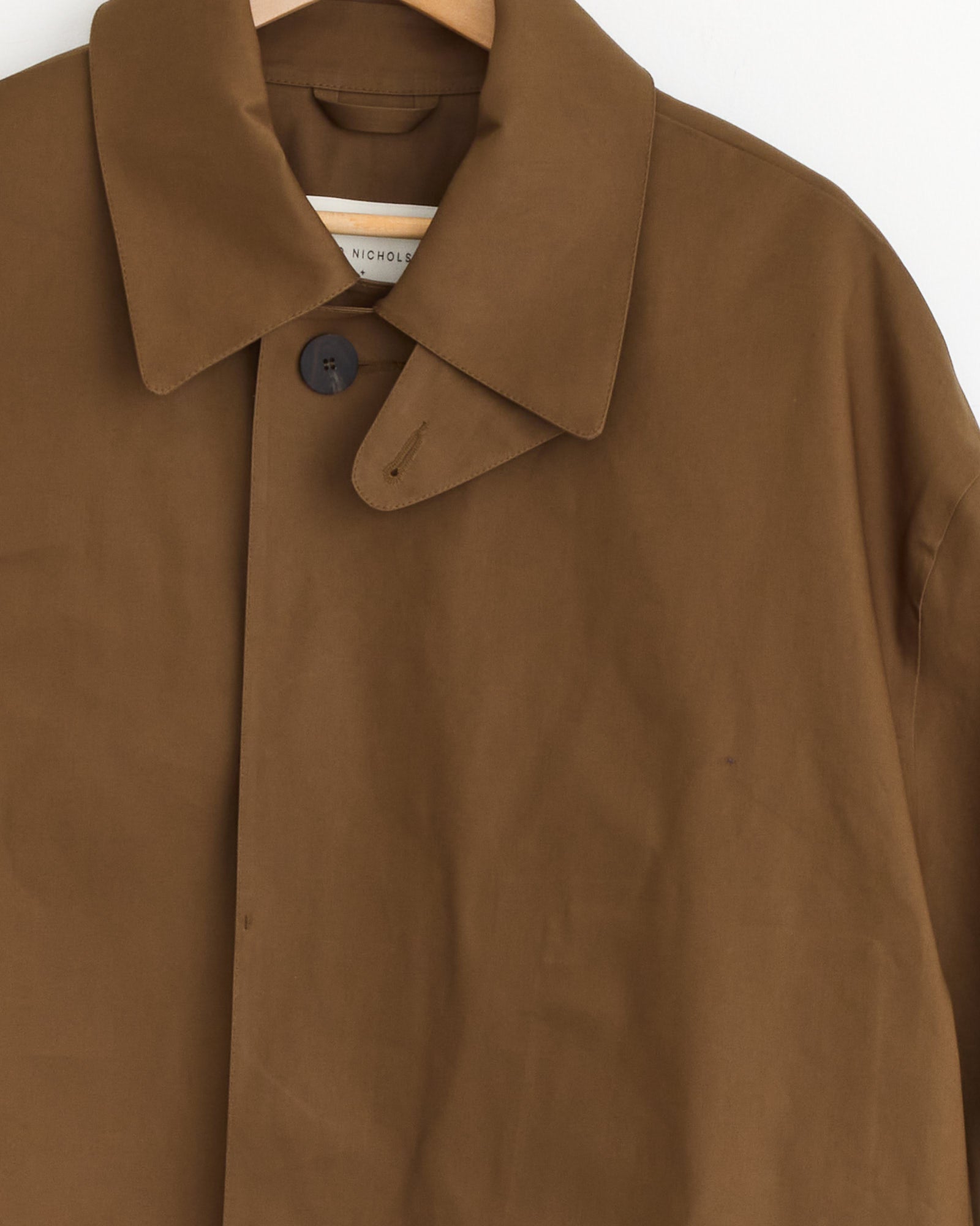 The Studio Nicholson x Mackintosh Ettrick Long Coat in Peat, featuring a pointed collar and two-button closure, hangs on a wooden hanger against a white background—a timeless brown waterproof raincoat.