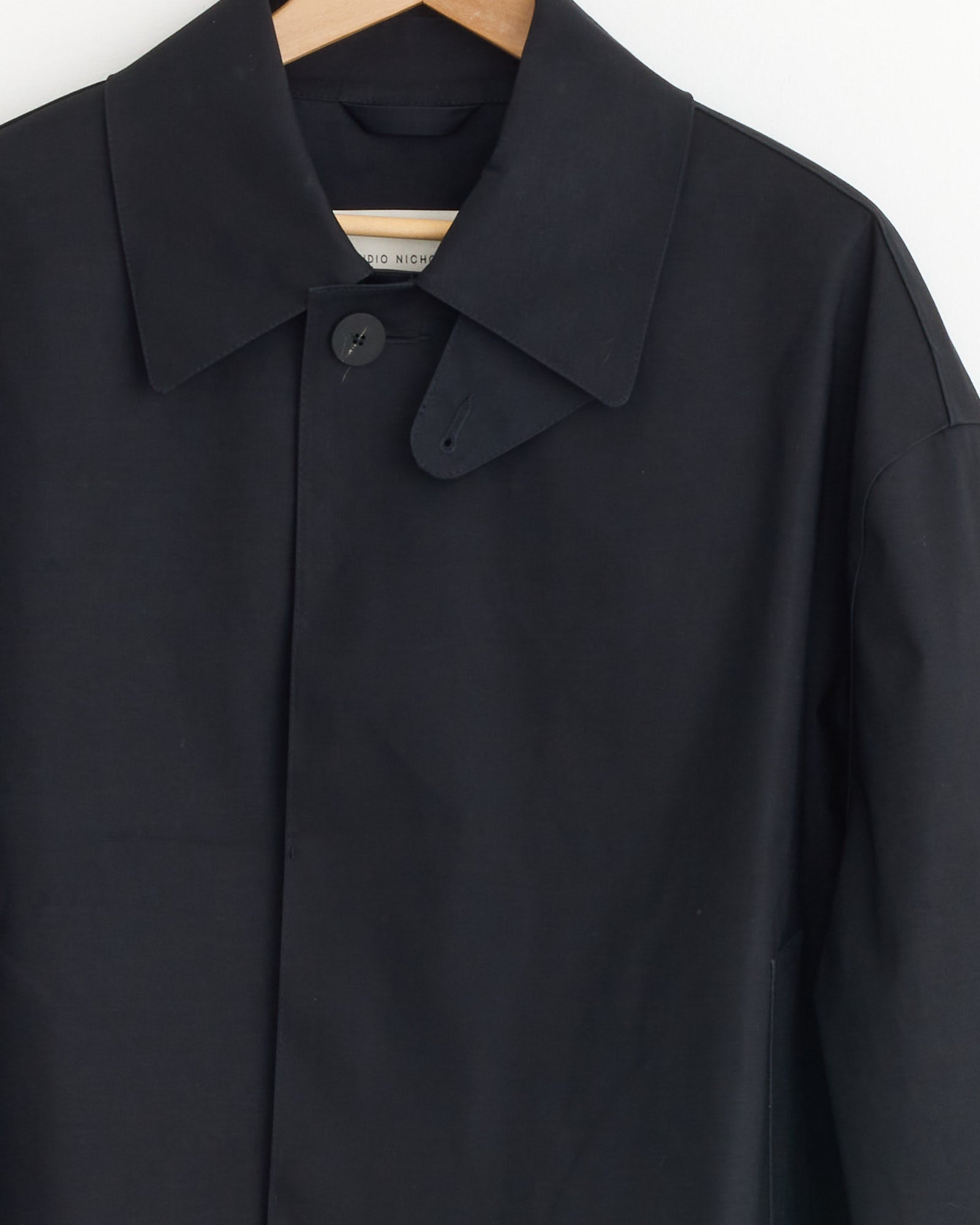 A close-up of the Studio Nicholson x Mackintosh Teviot Bonded Short Coat in Black shows its structured collar, minimalist design, and concealed button placket while hanging on a wooden hanger.