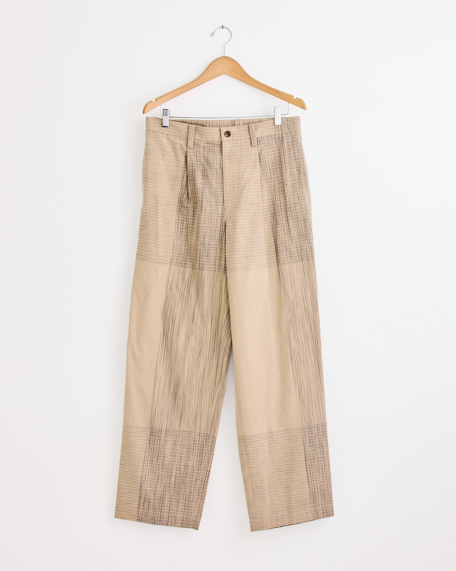 The Kasuri Pant in Beige by IM MEN features a beige and light brown plaid pattern with button and zip closure, shown on a wooden hanger against a white wall. For fit or sizing help, contact us via our online chat.
